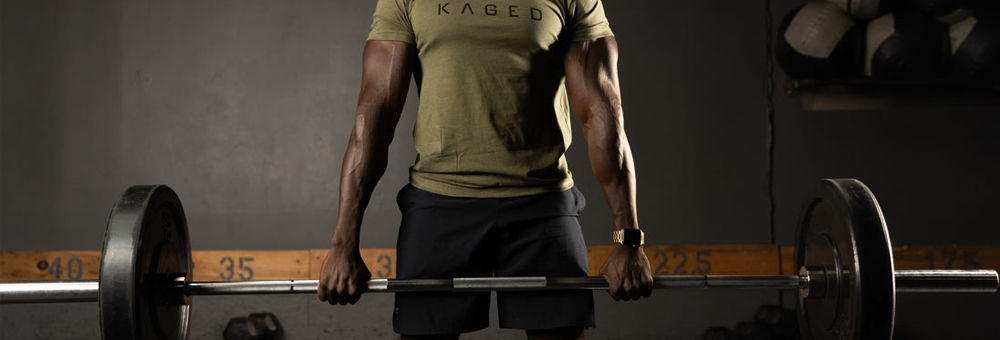 Person deadlifting a loaded barbell in a dim gym, wearing an olive KAGED t-shirt