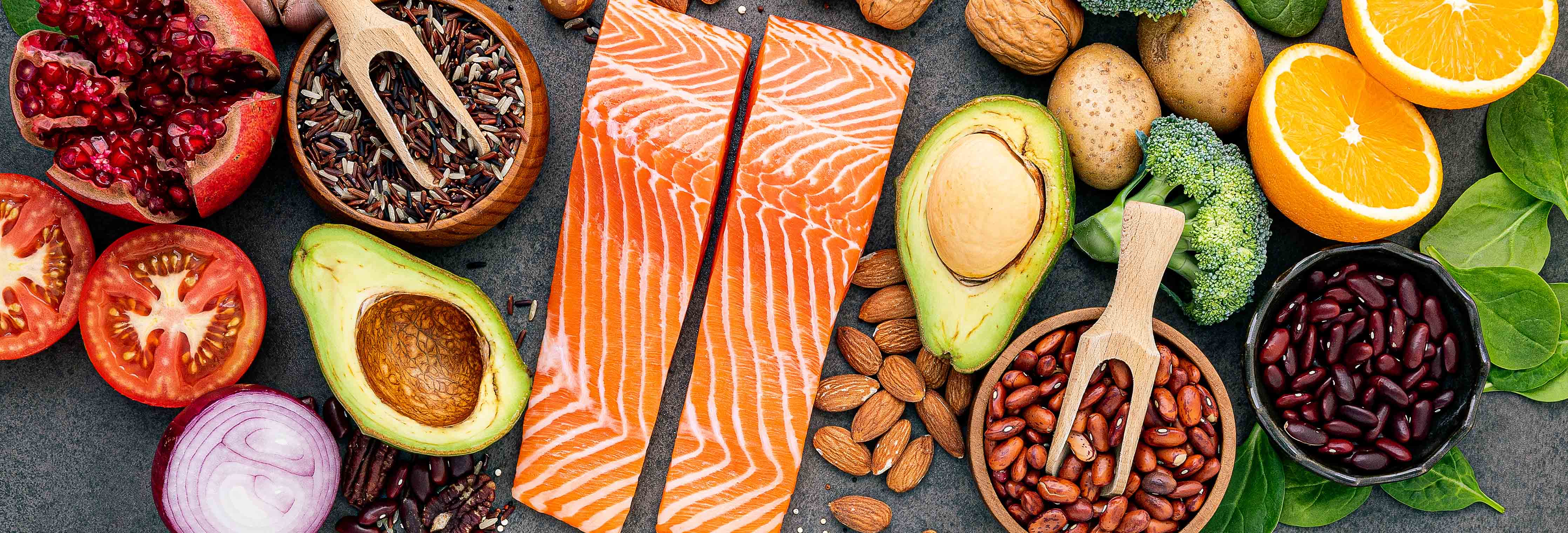 Assorted high-protein foods: raw salmon fillets, avocado halves, almonds, kidney beans, tomatoes, spinach and orange slices on a dark background