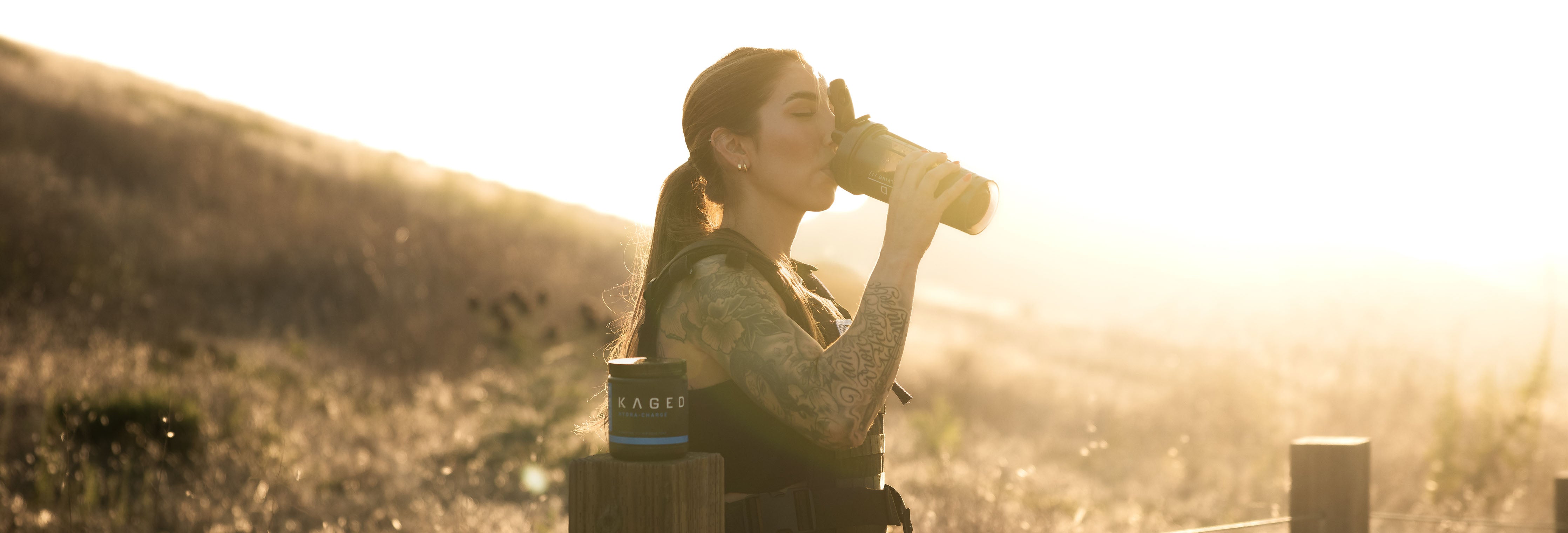 Woman drinking a peri-workout shake from a shaker bottle on a sunlit outdoor trail with a Kaged supplement tub on a wooden post