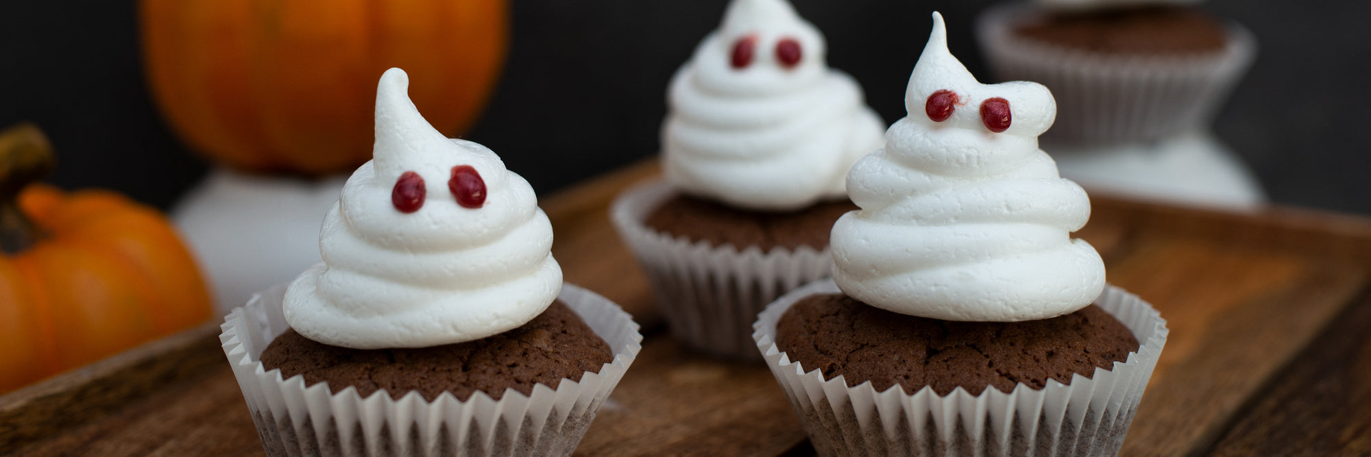 Front view of chocolate protein cupcakes with ghost-shaped white frosting and red candy eyes on a wooden board with pumpkins