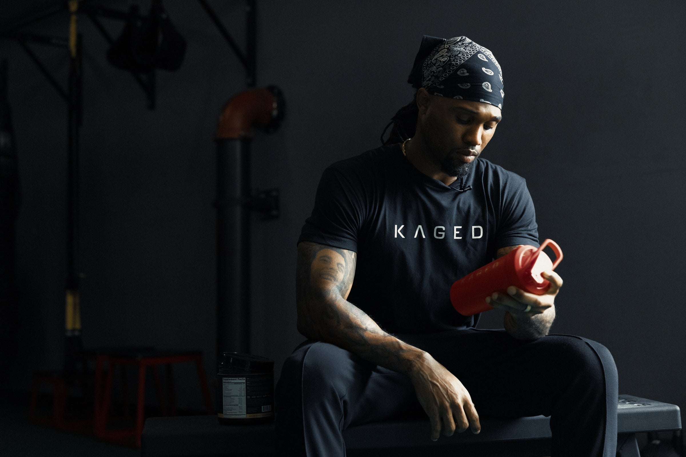 Man sitting on a gym bench inspecting a red shaker bottle while wearing a black KAGED t-shirt