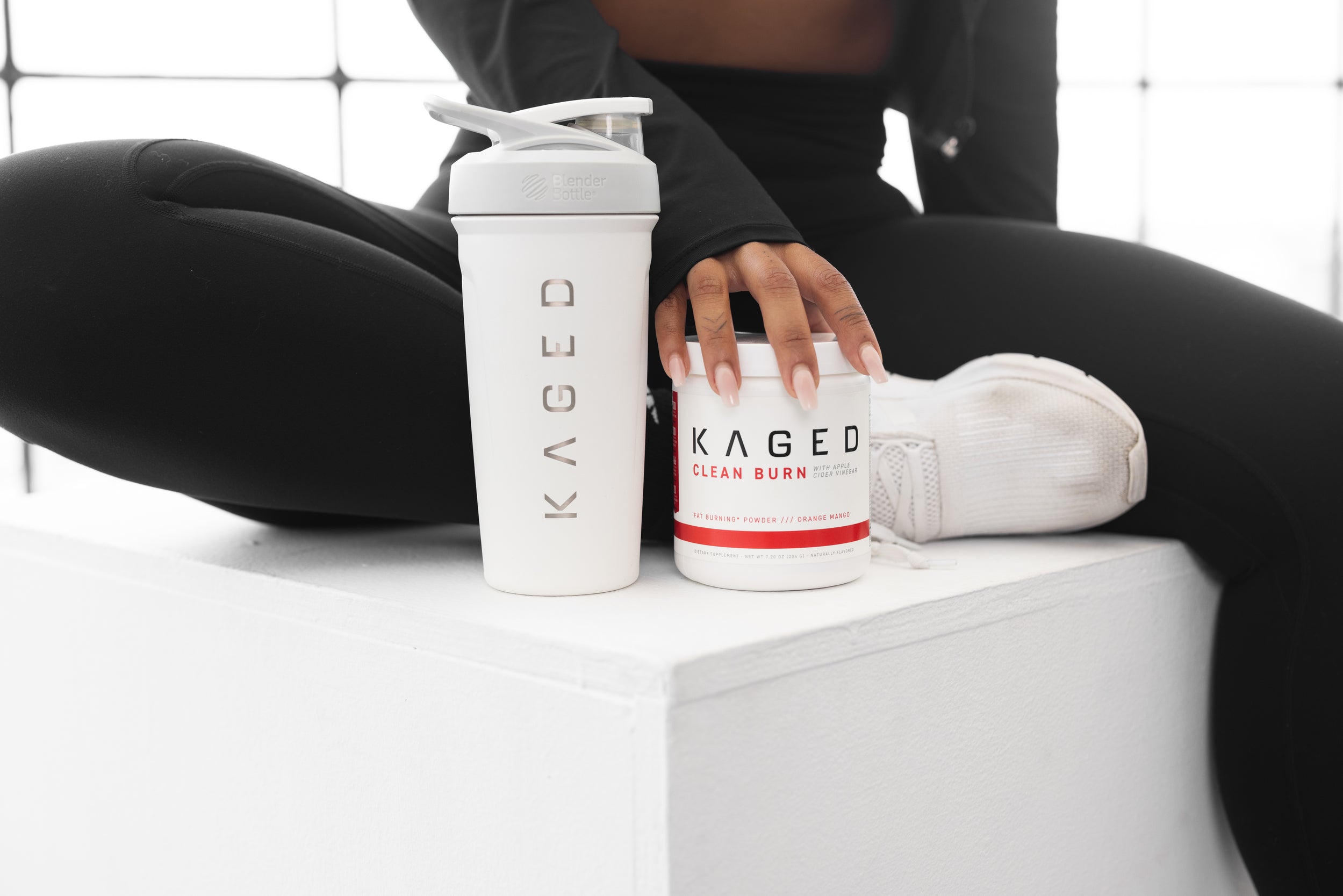 Front view of KAGED Clean Burn tub and matching white KAGED shaker, model hand and black leggings on white block