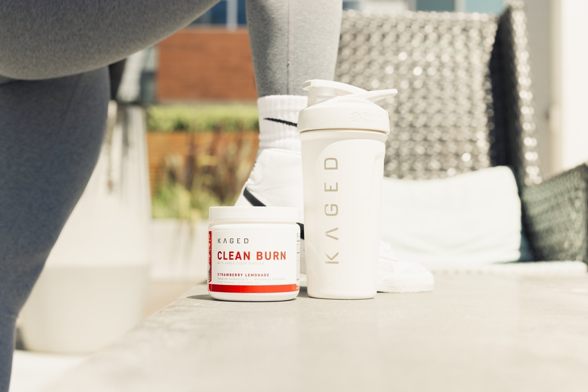Front view of KAGED Clean Burn strawberry lemonade tub and white KAGED shaker on bench with person in leggings and sneakers on patio