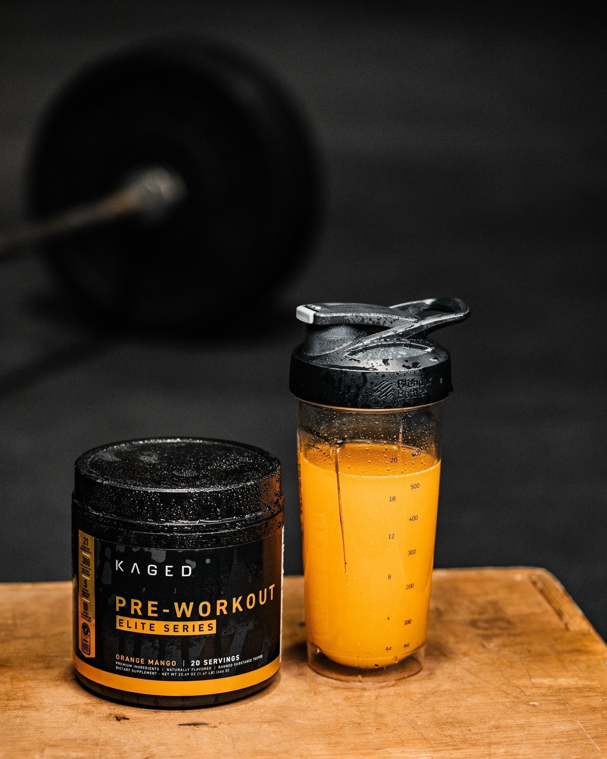 Front view of KAGED Pre-Workout tub and a shaker with orange-mango drink on a wooden bench, blurred barbell in background