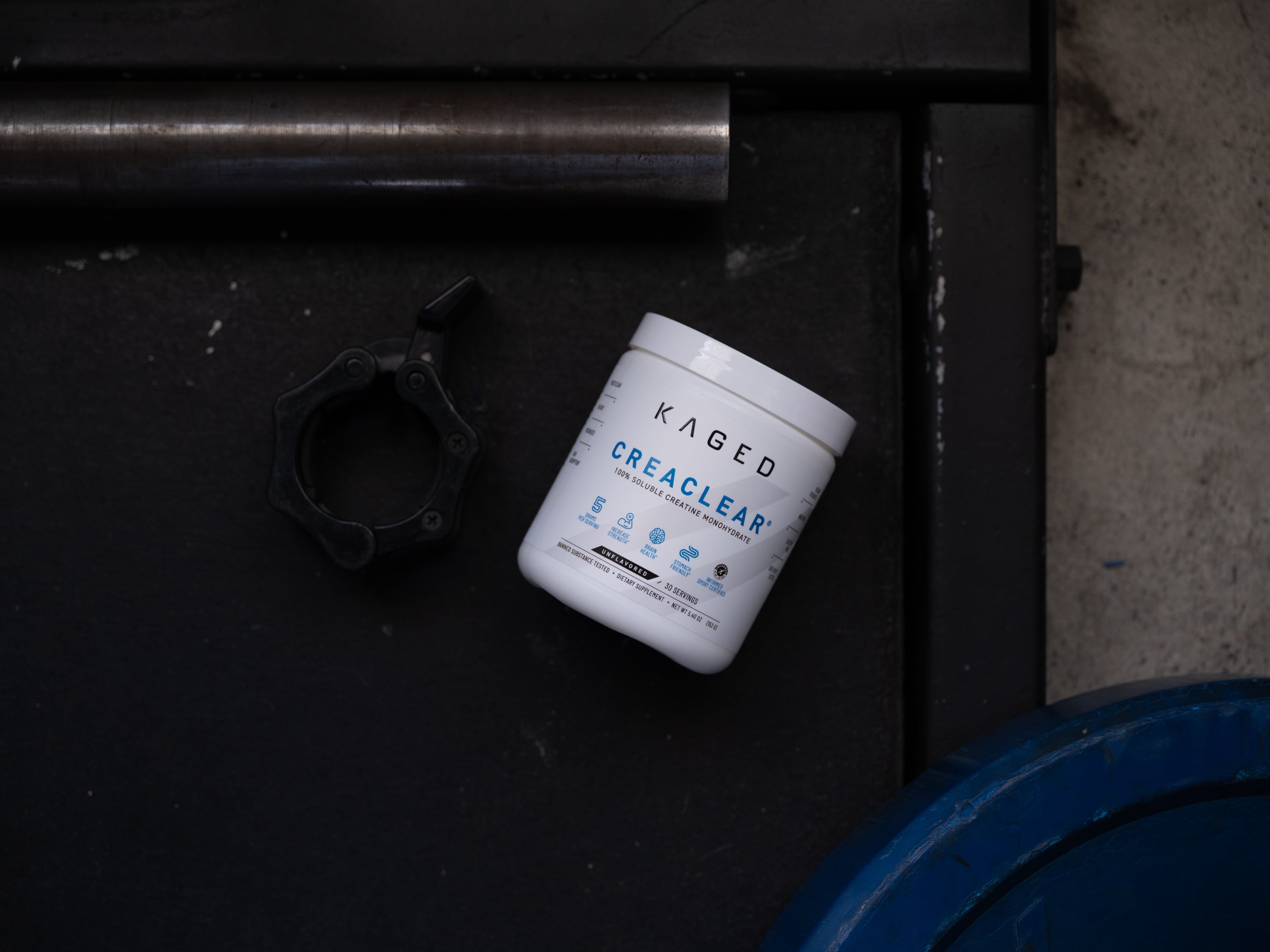 Angled view of KAGED CreaClear creatine tub on a dark gym floor beside a barbell collar and blue weight plate