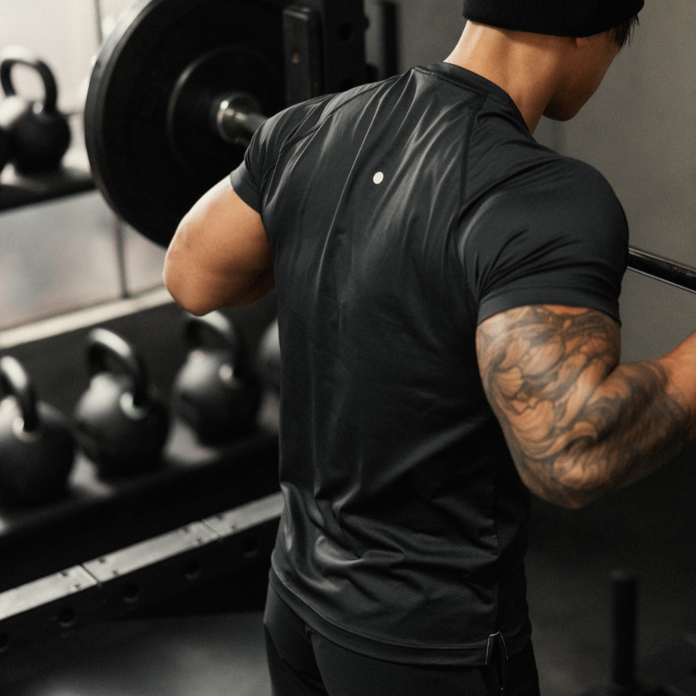 Rear view of Ascent Performance Tee in black on model showing athletic slim fit and small reflective back logo in a gym
