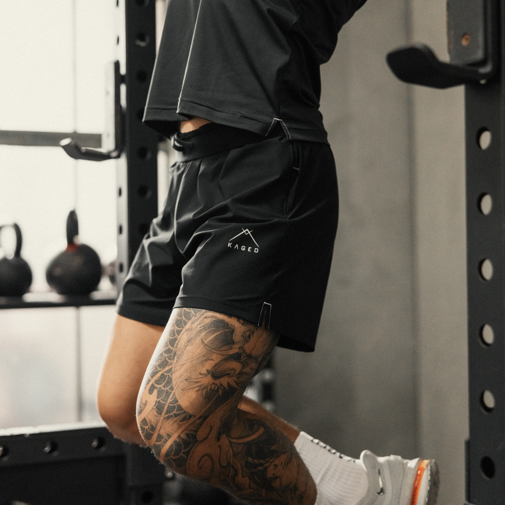 Side view of Ascent Performance Shorts in black on model showcasing athletic fit and KAGED logo in a gym setting