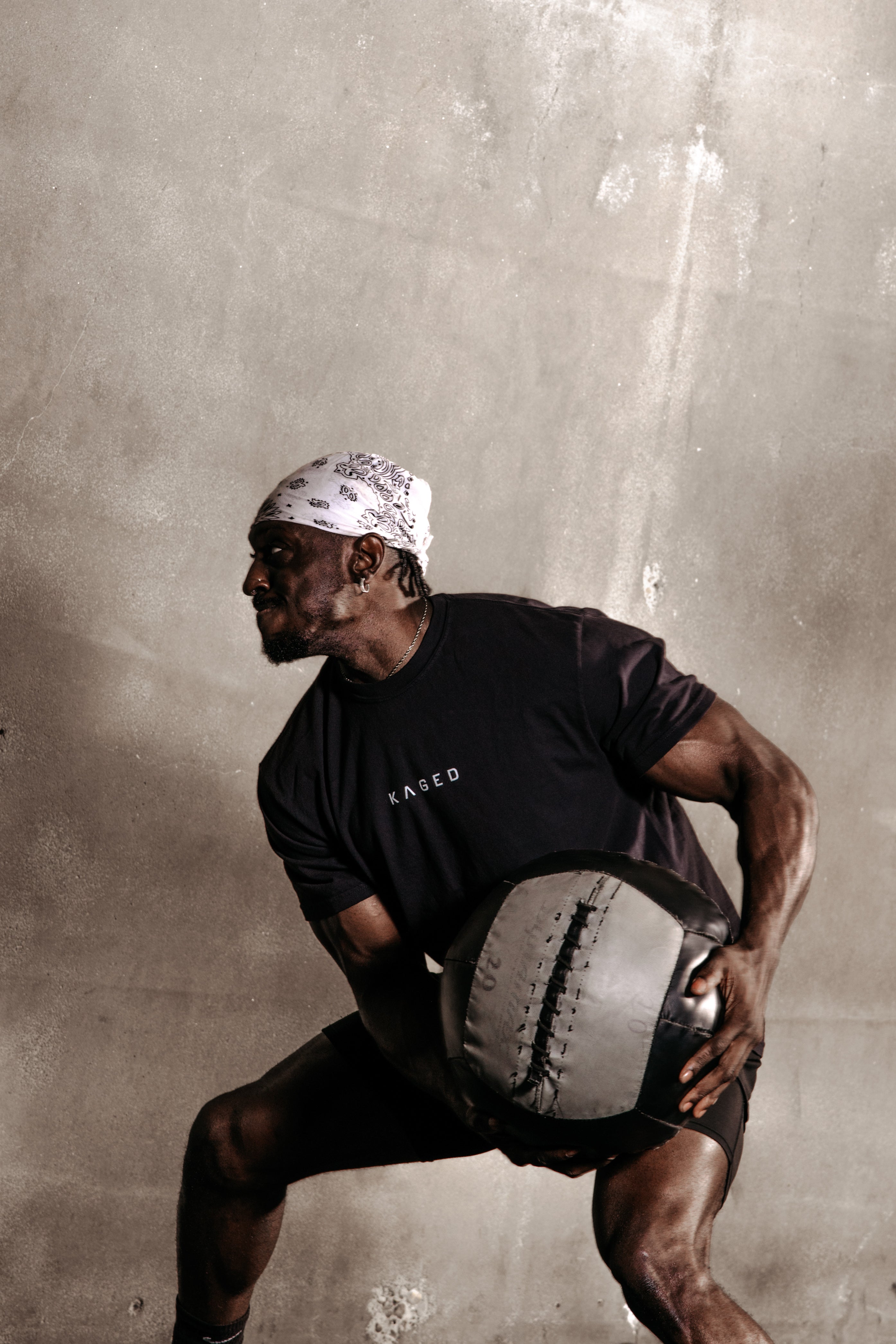 Muscular man twisting with a medicine ball in front of a concrete wall, wearing a KAGED t-shirt and white bandana