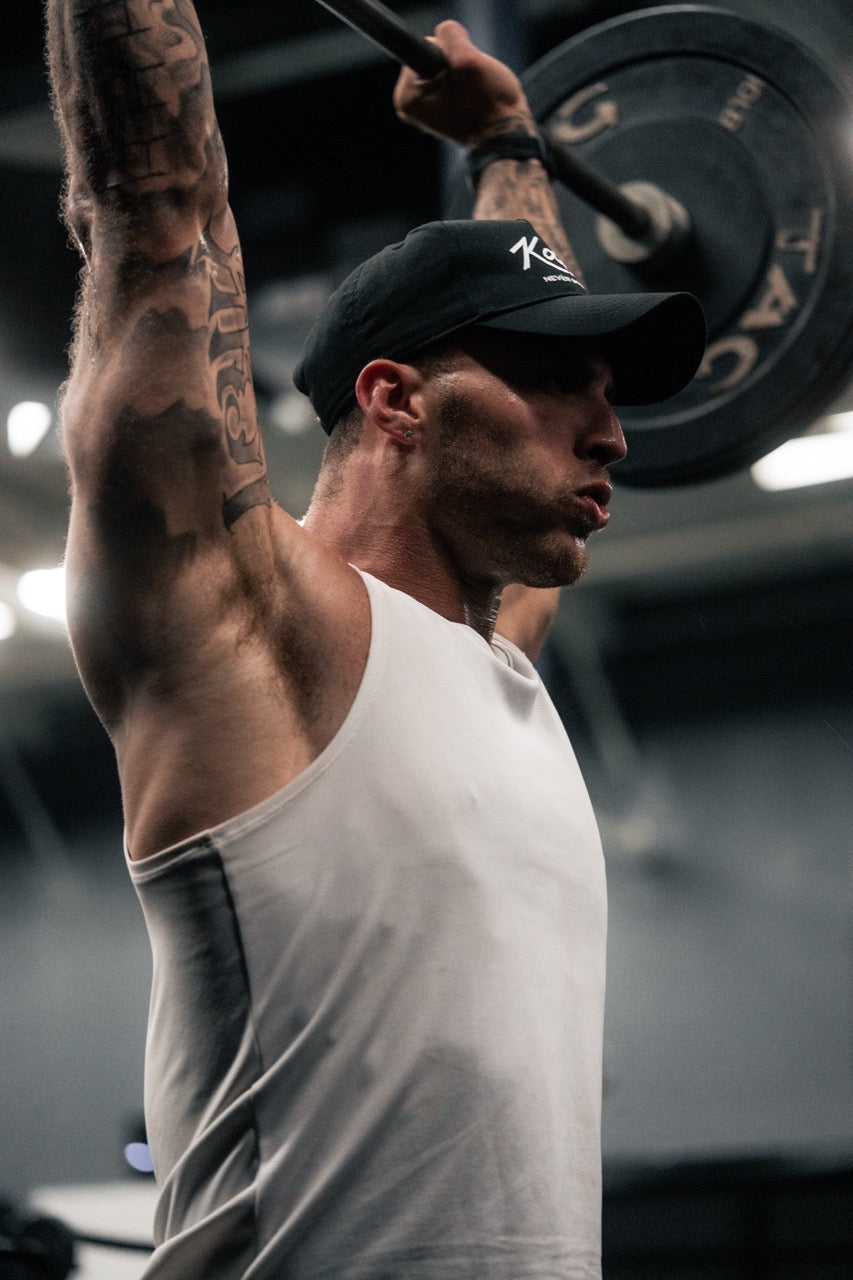 Athlete performing an overhead barbell press in a gym, wearing a white tank top and black cap, muscles tensed