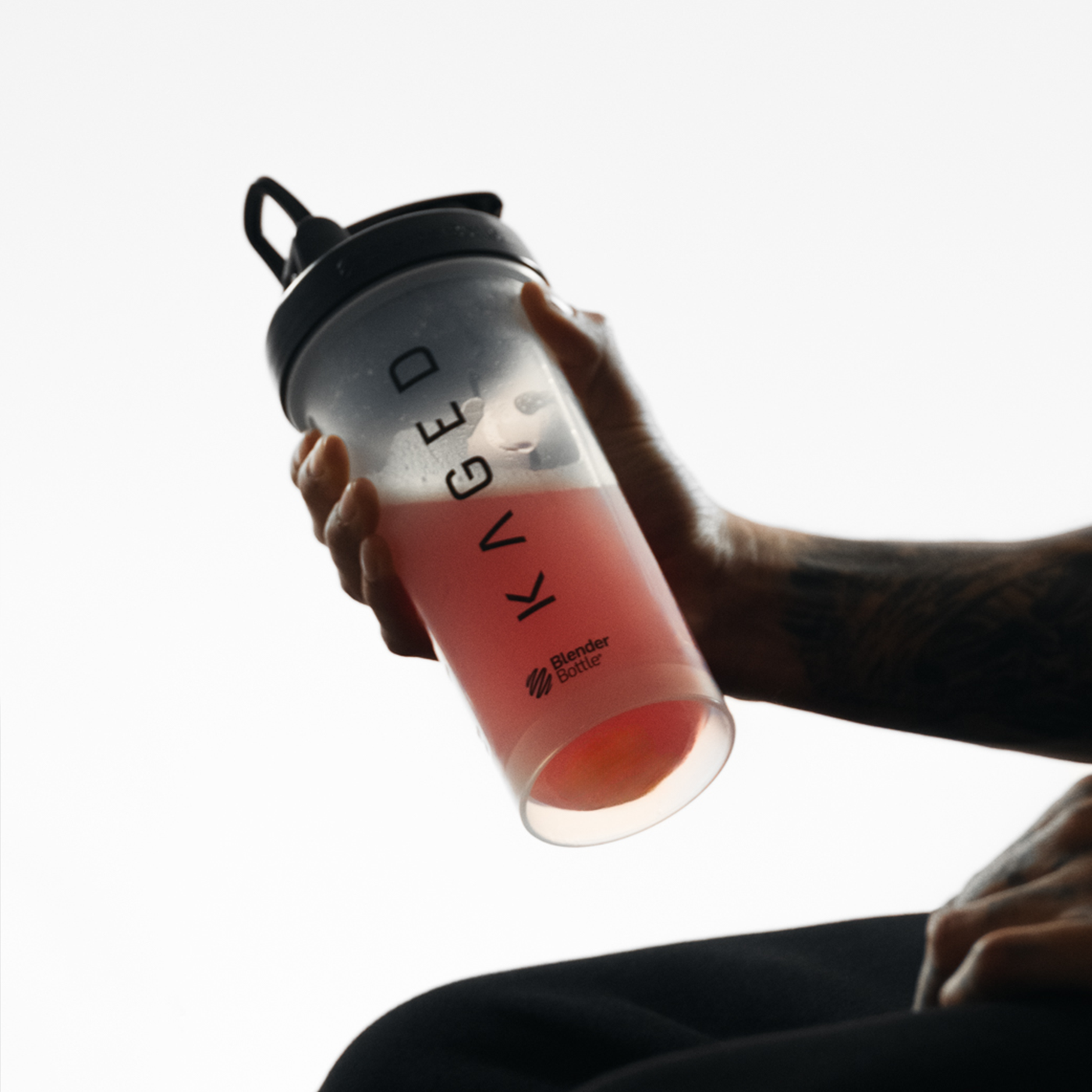 Tilted view of Blender Bottle - Clear held in hand with black SpoutGuard lid, carry loop, KAGED logo, and pink drink