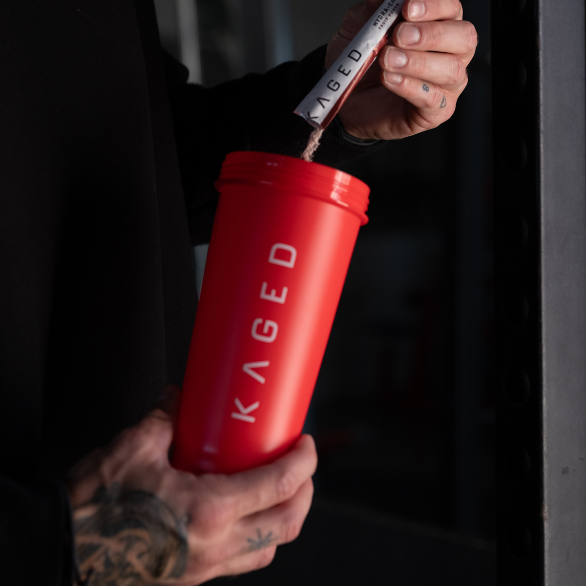 Person pouring supplement powder into crimson shaker bottle at a gym