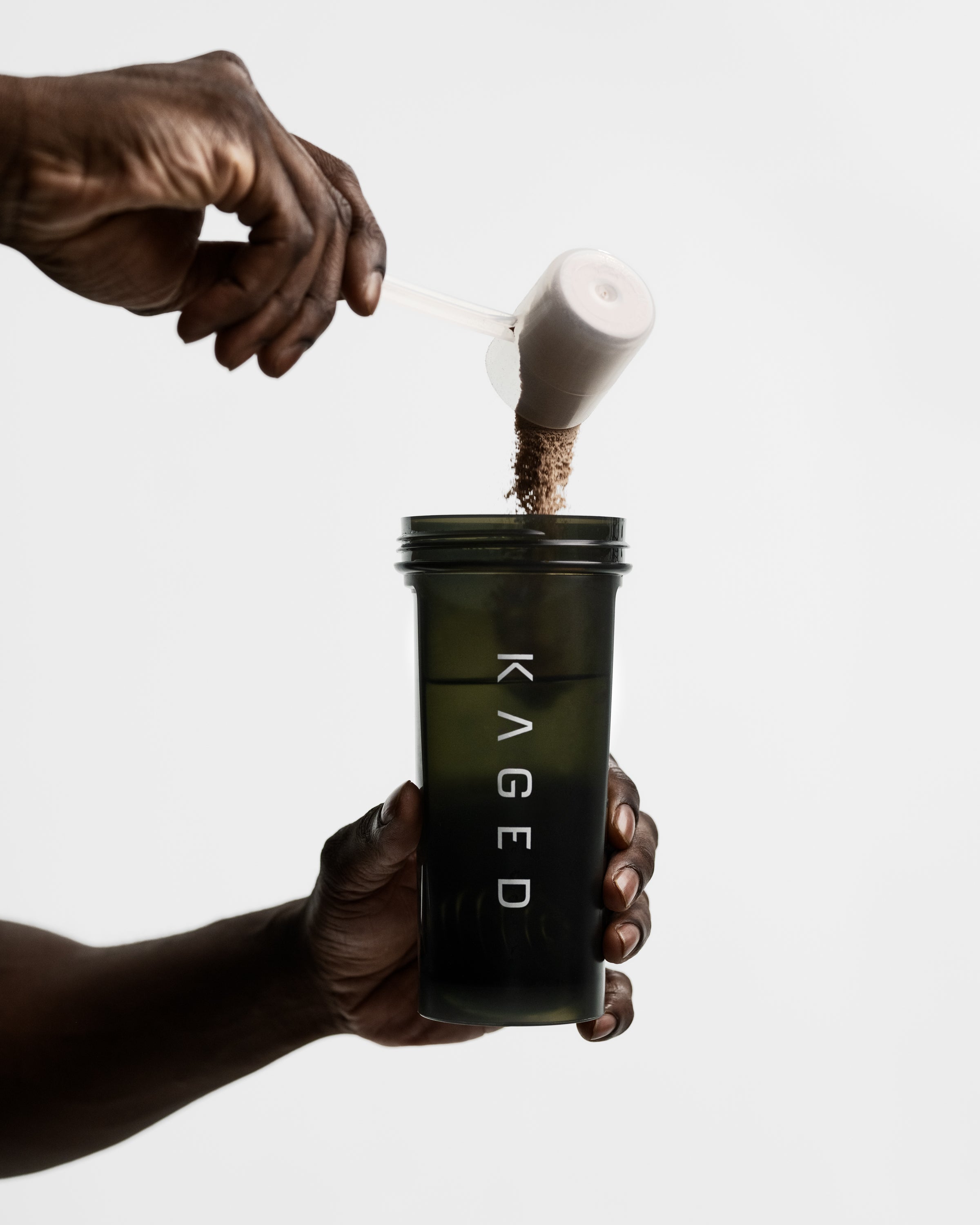 Hand pouring Clean Meal powder from a scoop into a dark green KAGED shaker bottle on a white background