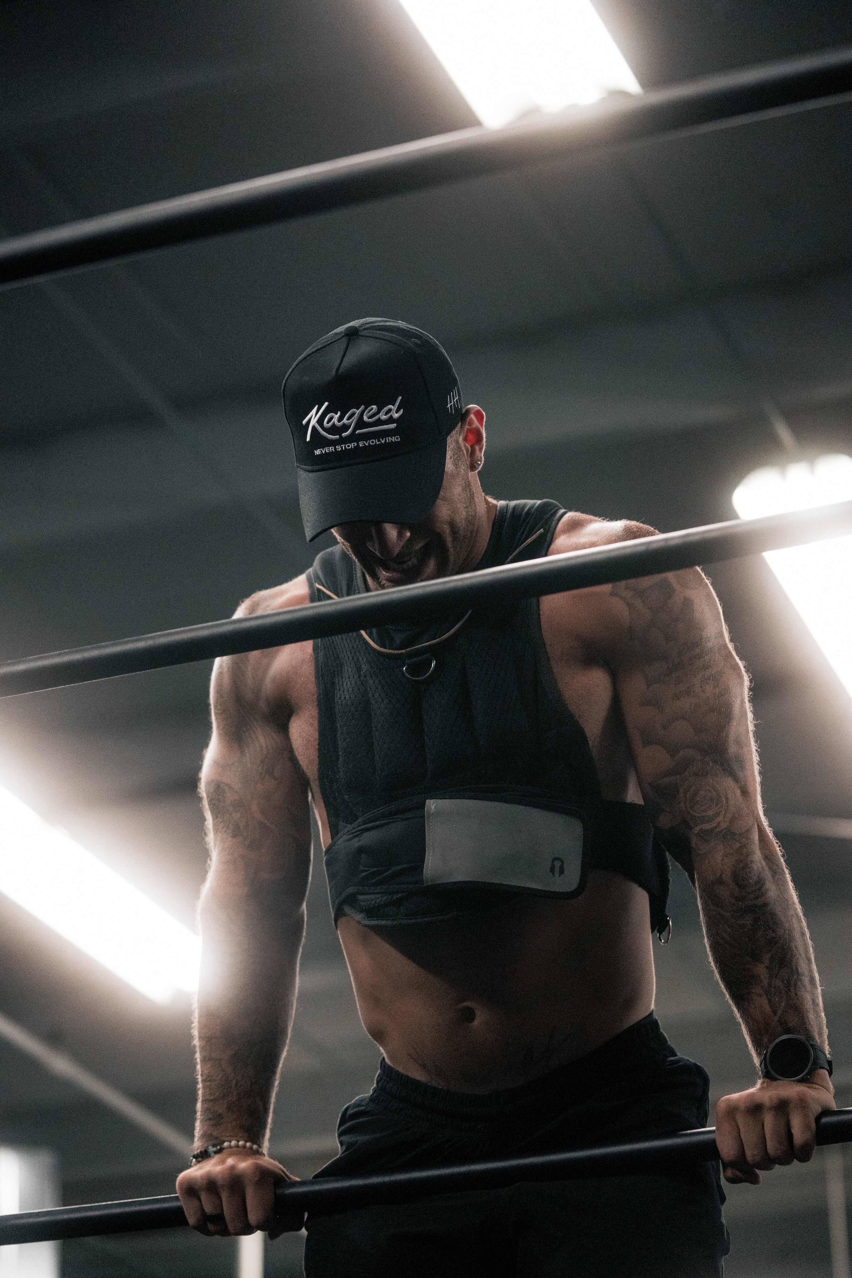 Man wearing Kaged cap and weighted vest doing dips on parallel bars in a dimly lit gym