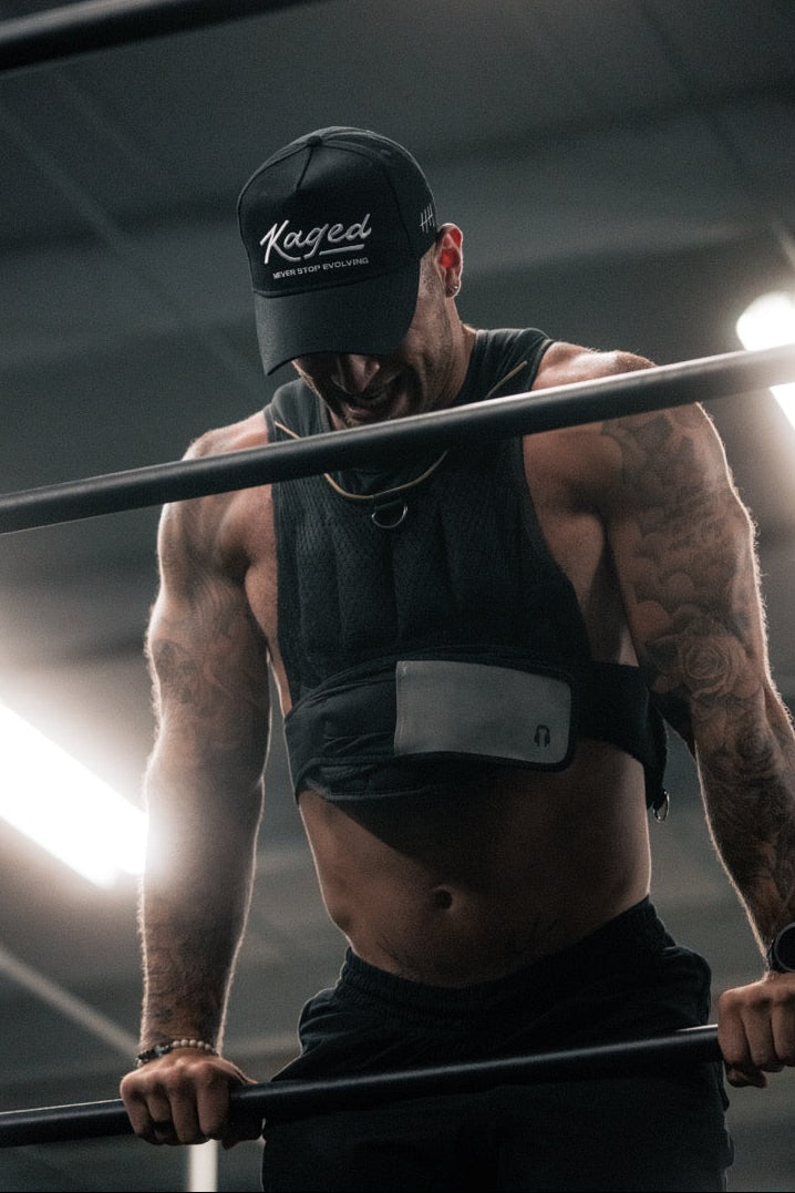Man doing parallel-bar dips in a gym, wearing a black Kaged cap and a training vest