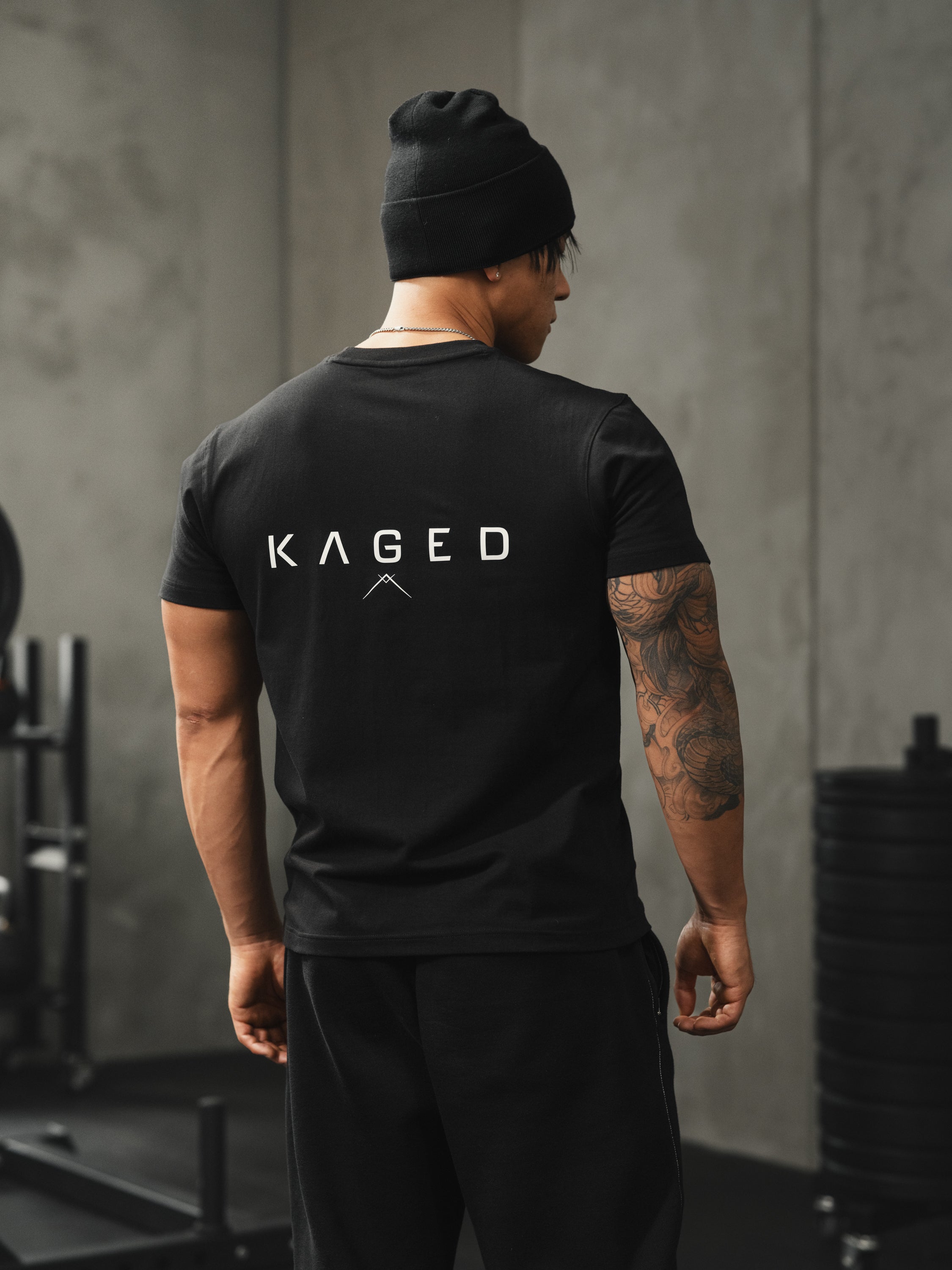 Rear view of black Ascent Tee on model showcasing large KAGED wordmark on back and fitted silhouette in a gym setting
