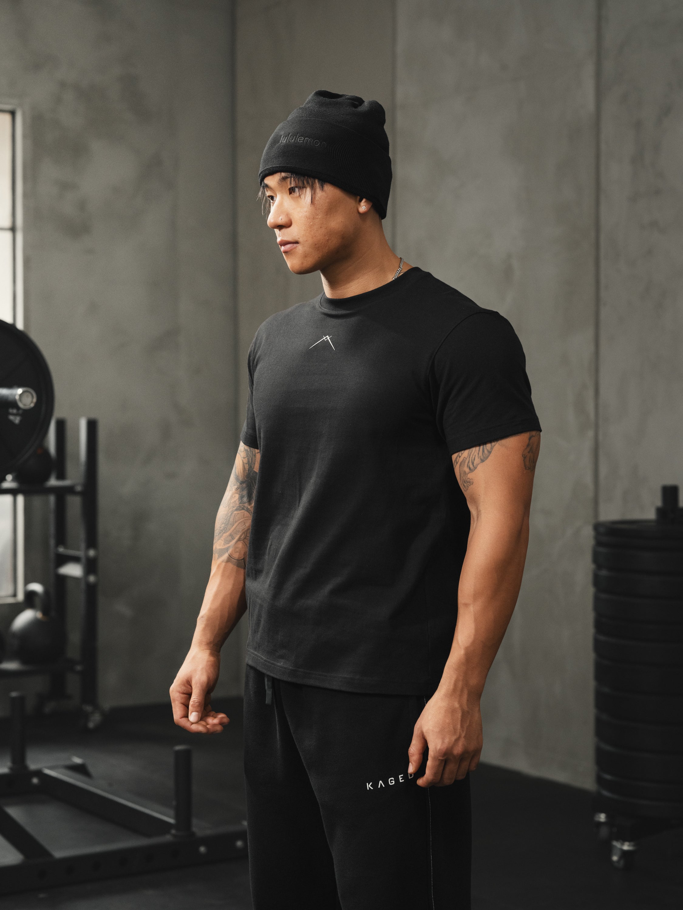 Model wearing fitted black Ascent tee standing in a gym with weight rack and plates in the background