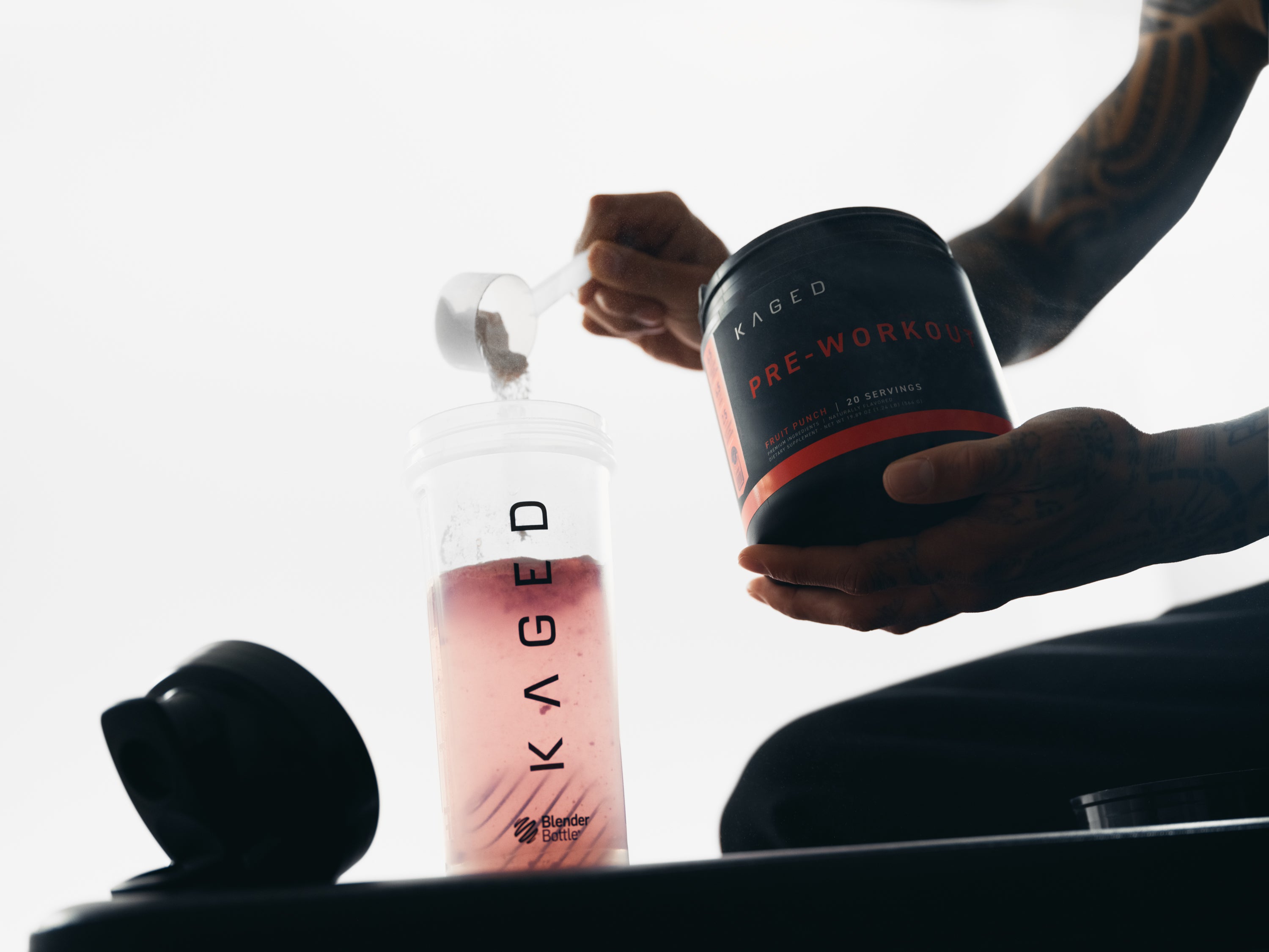 Person pouring KAGED pre workout powder into a clear KAGED shaker bottle with pink drink in a studio setting
