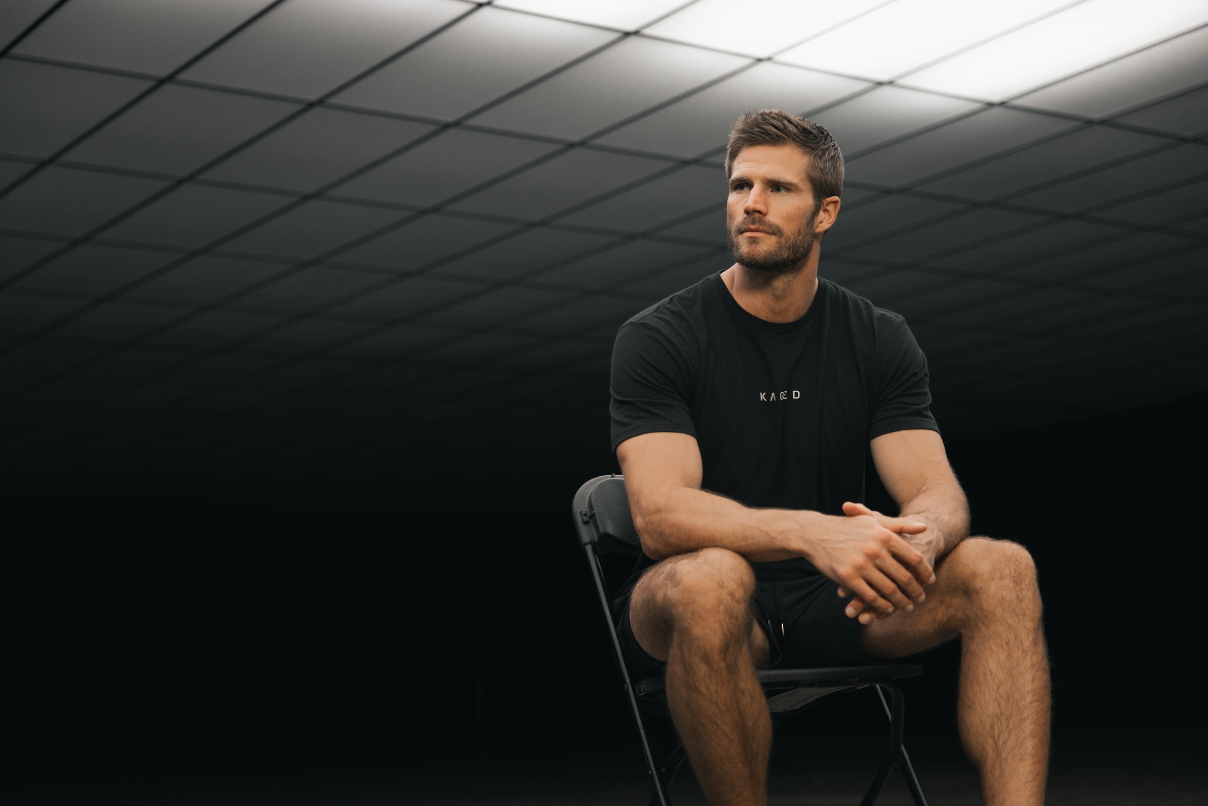 Man sitting on a chair wearing a black KAGED T-shirt with logo, looking to the side against a dark geometric ceiling background