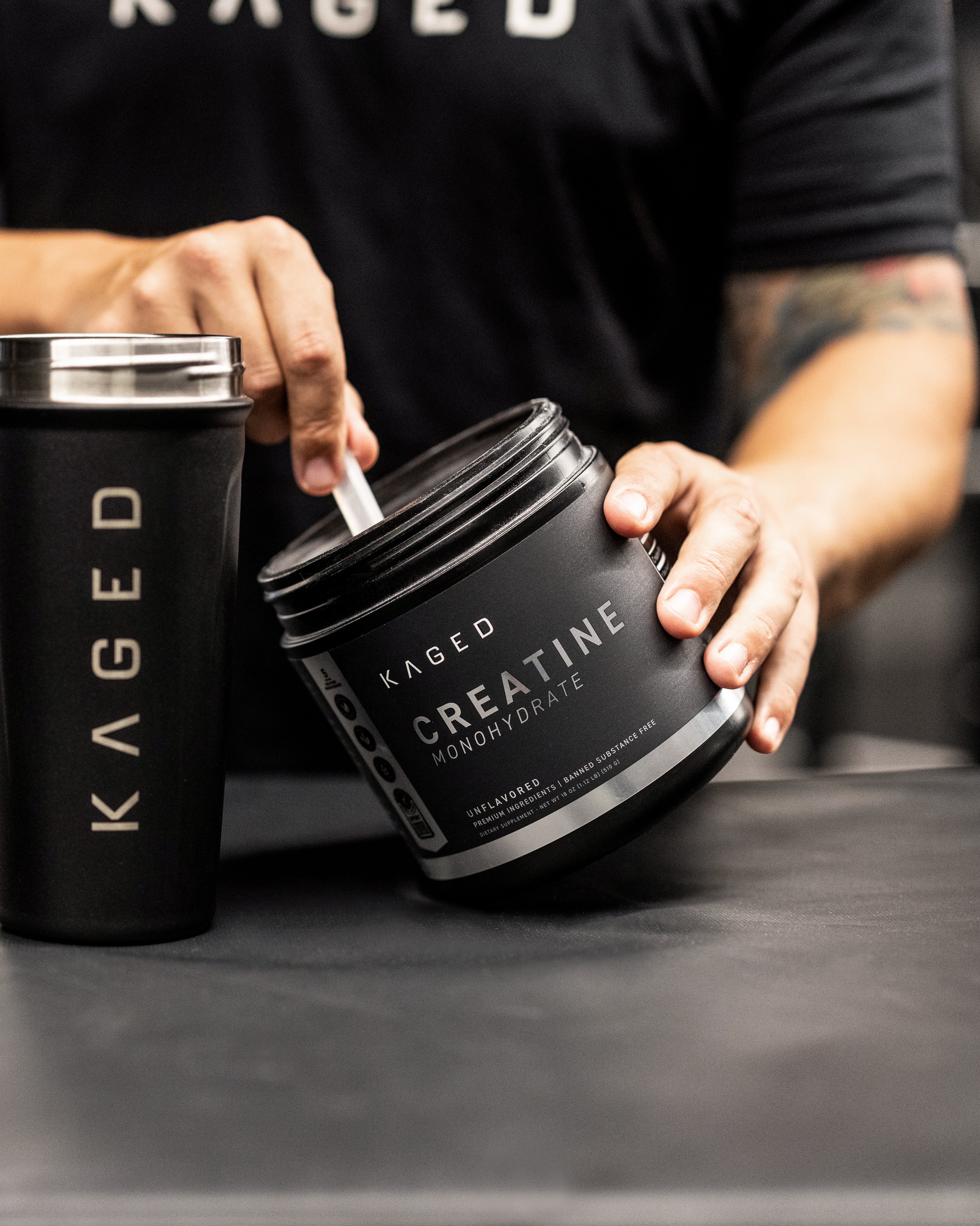 Hands scooping Kaged Creatine Monohydrate powder from a black tub next to a Kaged shaker