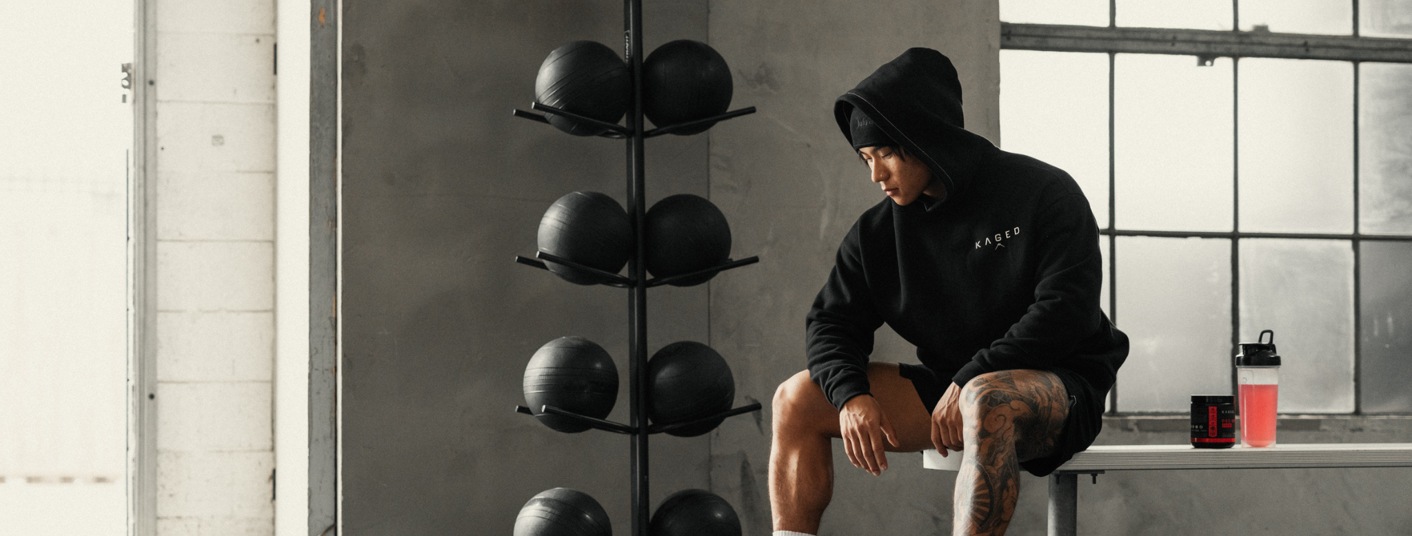 Model wearing a black KAGED hoodie sitting on a gym bench beside a supplement tub and a pink shaker bottle, medicine balls and industrial windows in the background