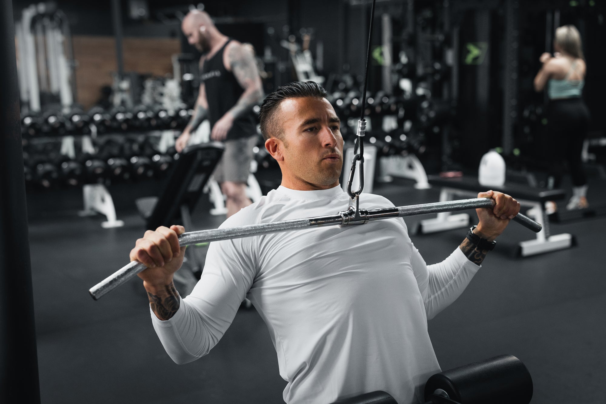 Man performing a seated lat pulldown on a cable machine in a gym, wearing a white long-sleeve shirt