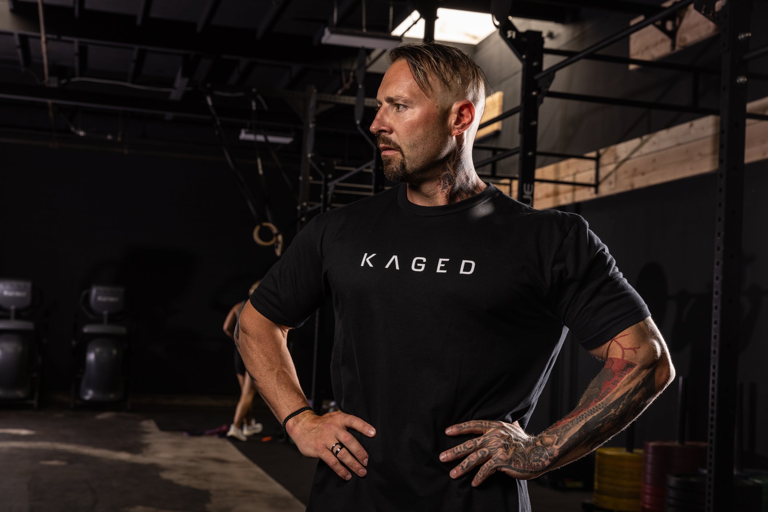Tattooed man standing with hands on hips in a dim gym wearing a black shirt with large KAGED text