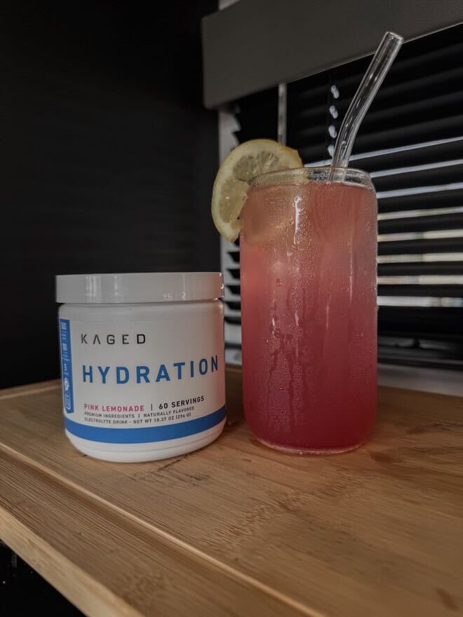 Front view of KAGED Hydration Pink Lemonade tub beside a pink mocktail in a glass with lemon slice on a wooden surface