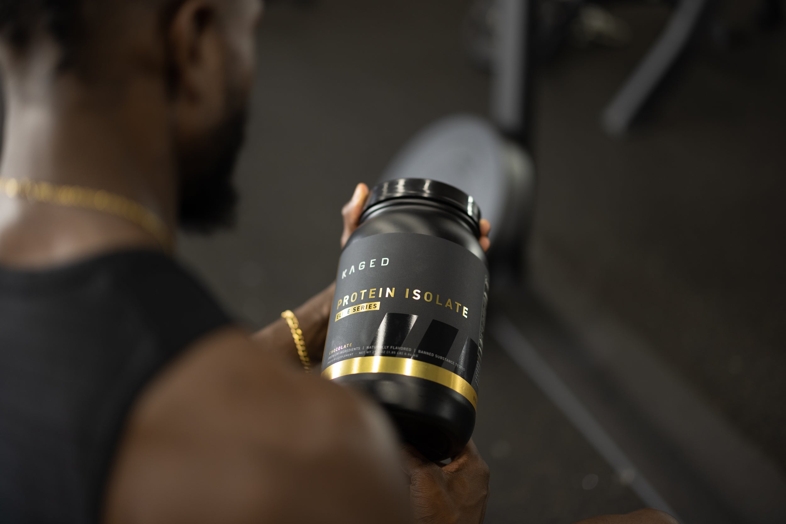Angled view of KAGED Protein Isolate tub with black and gold label held by a person in a gym setting
