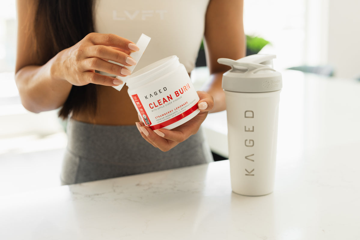 Waist-level close-up of Kaged Clean Burn supplement tub held open to show the label 'Clean Burn Strawberry Lemonade' with a white Kaged shaker on a bright kitchen counter