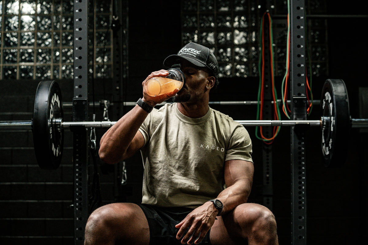 Man drinking an orange pre workout shake while seated in front of a loaded barbell rack in a gym