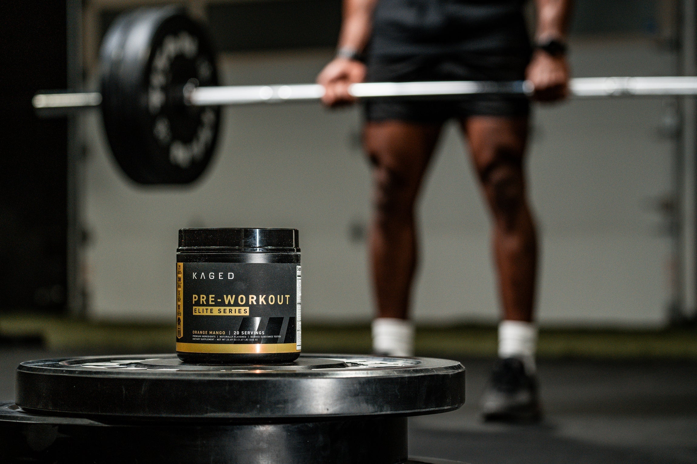 Three-quarter view of KAGED Pre-Workout tub showing PRE-WORKOUT label and serving info with a blurred weightlifter deadlifting in a gym