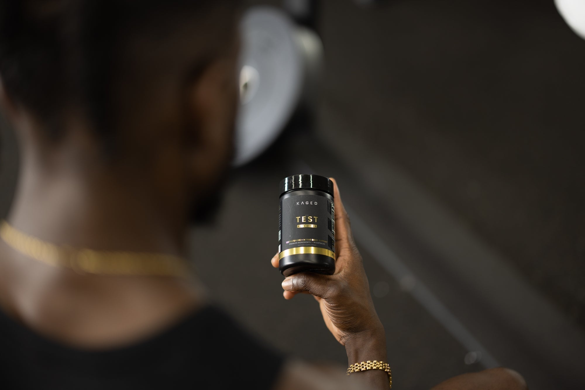 Hand holding KAGED TEST supplement jar with black-and-gold label in a gym with blurred weights in background