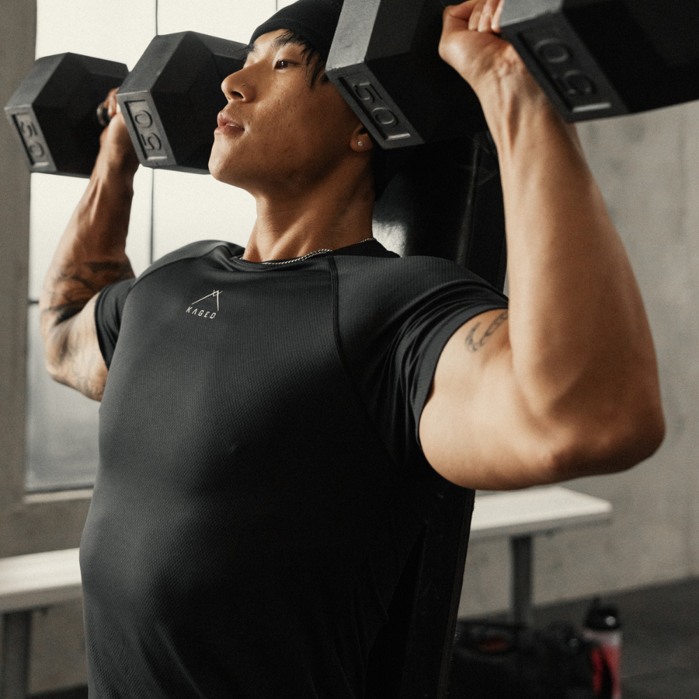 Male athlete performing seated dumbbell shoulder press wearing black athletic-fit Ascent Performance Tee in gym
