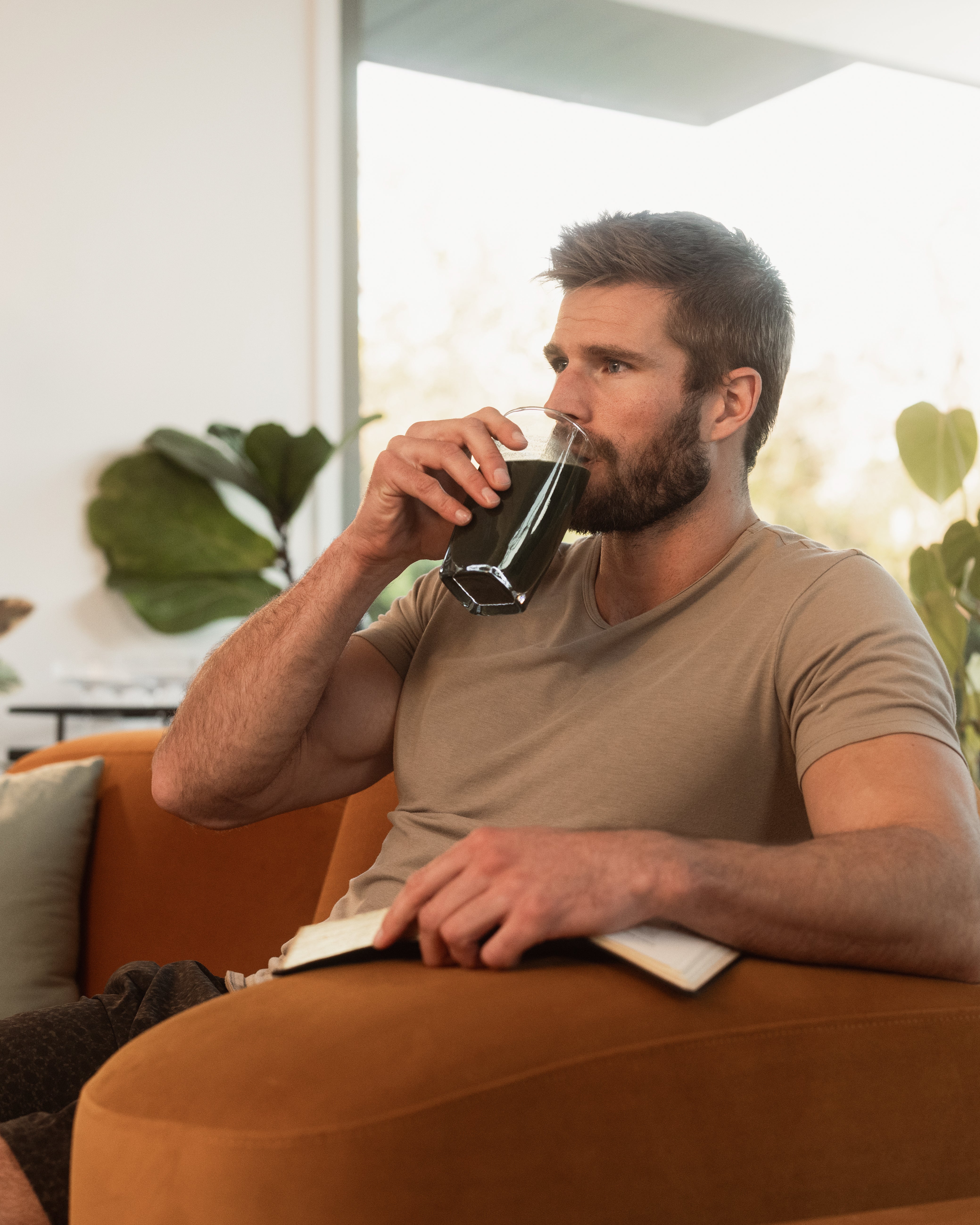 Man sipping a glass of green superfood supplement on an orange couch at home while holding a notebook