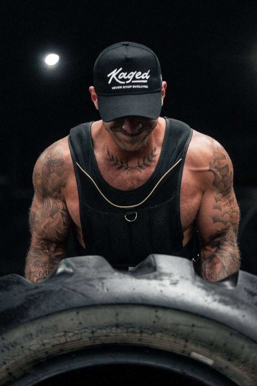 Man flipping a large tire in a dim gym wearing a Kaged cap and weighted vest