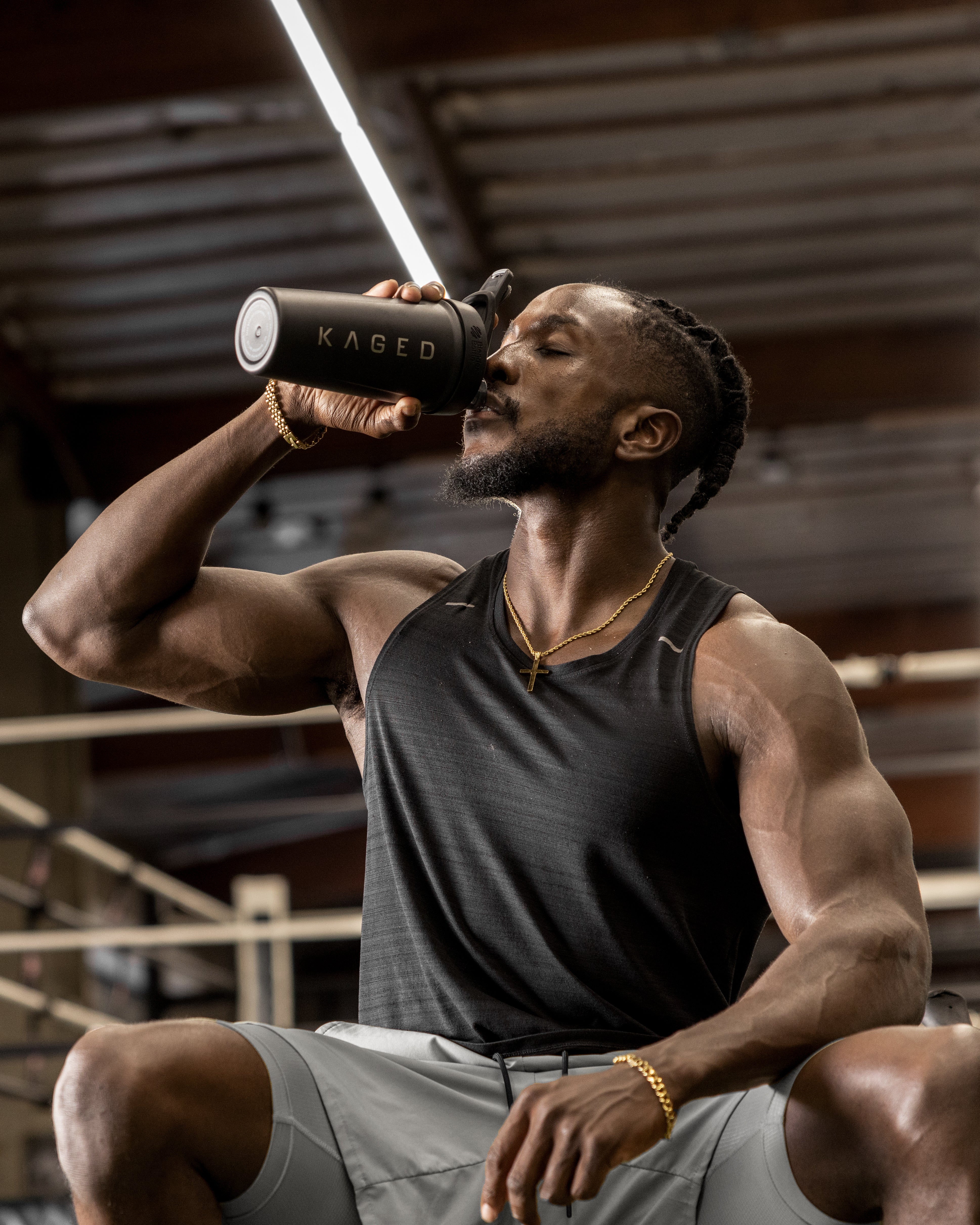 Muscular man drinking from a black bottle labeled KAGED in a gym