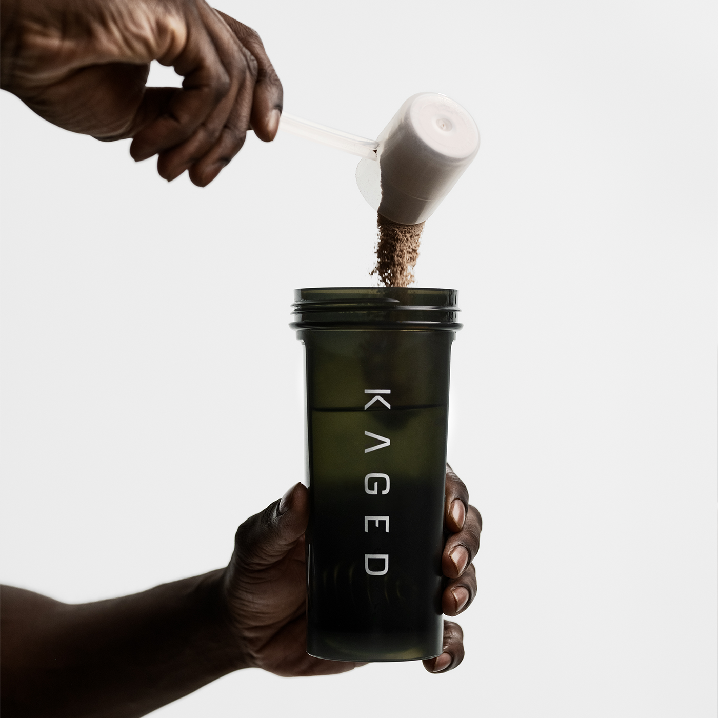 Hands pouring a scoop of brown supplement powder into a dark KAGED BlenderBottle Classic 28 oz against a white background