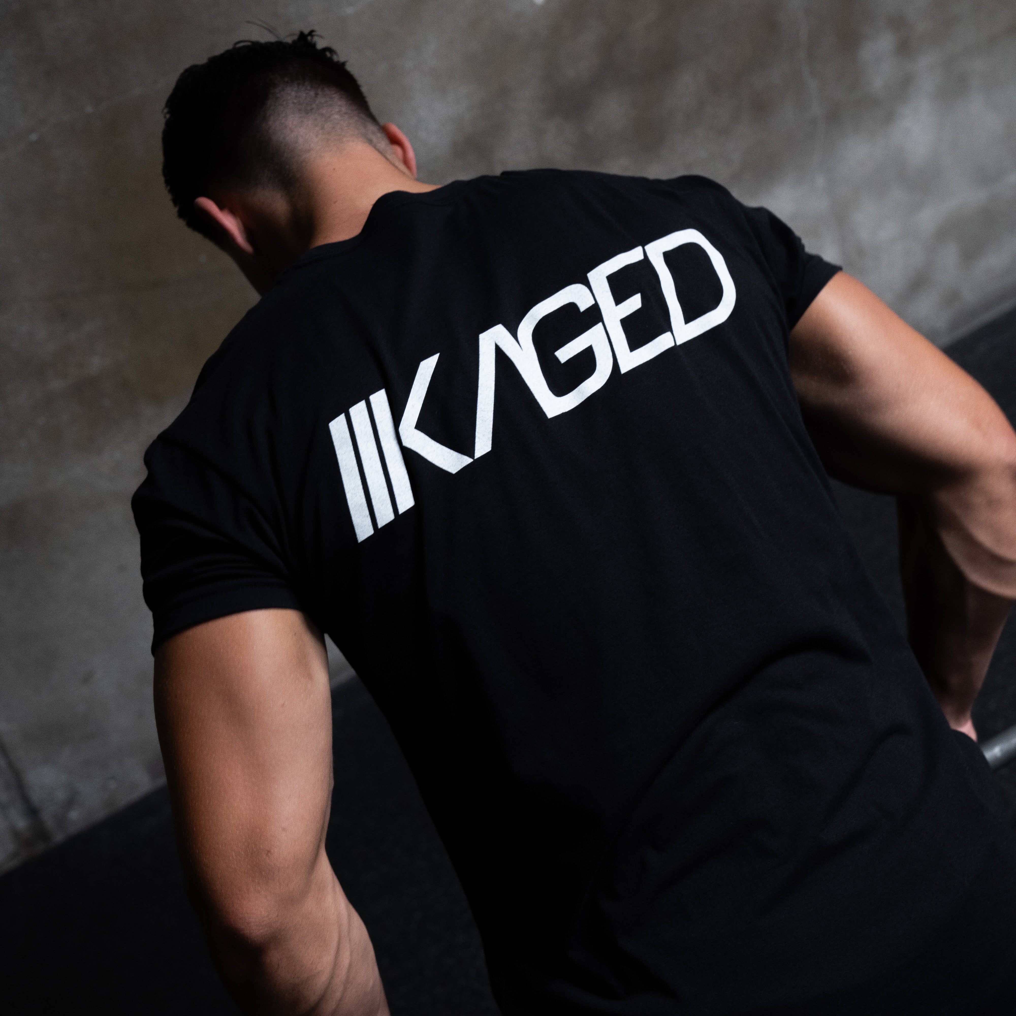 Rear view of Slant Tee in black showcasing large white angled KAGED logo on the back with gray concrete background #color_black