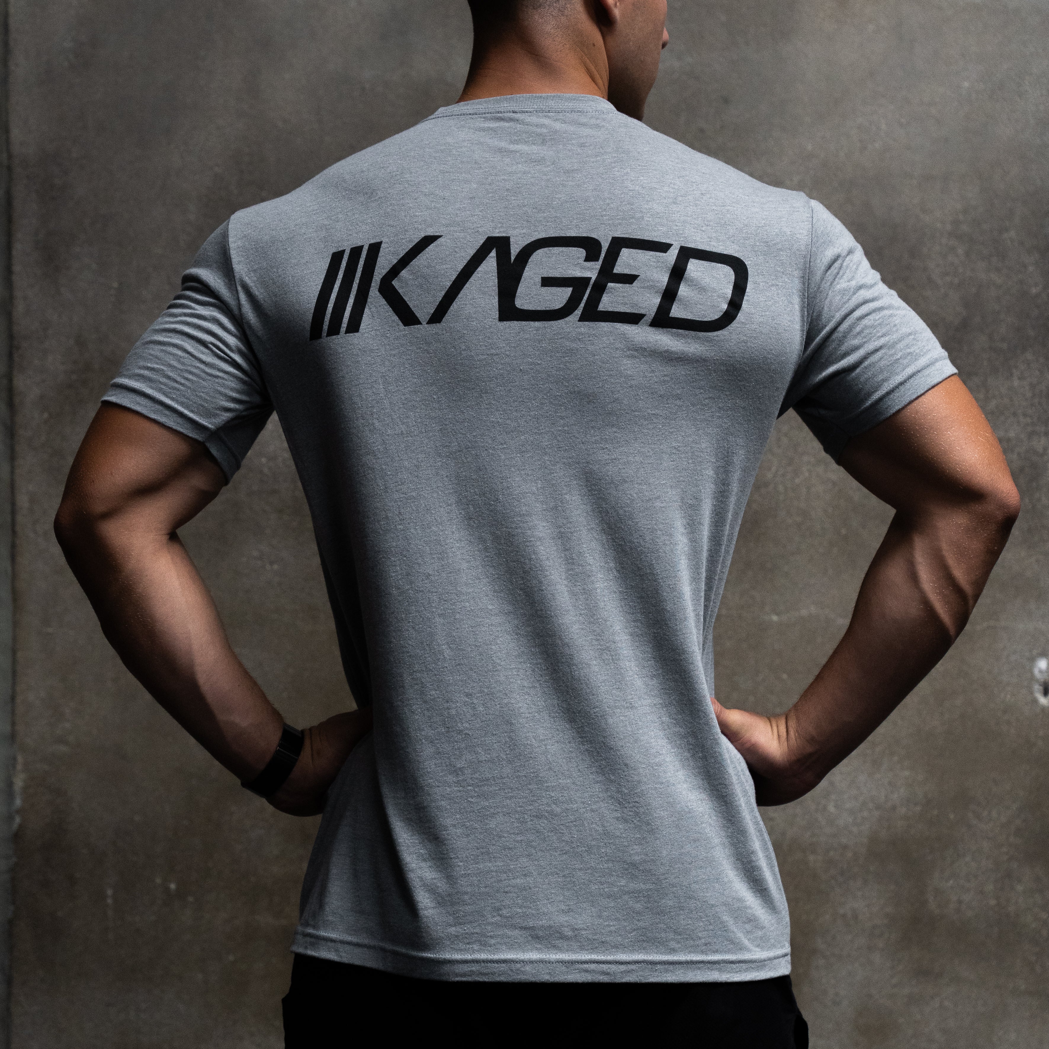 Back view of Slant Tee in gray showcasing large black KAGED logo across upper back with concrete wall background #color_gray