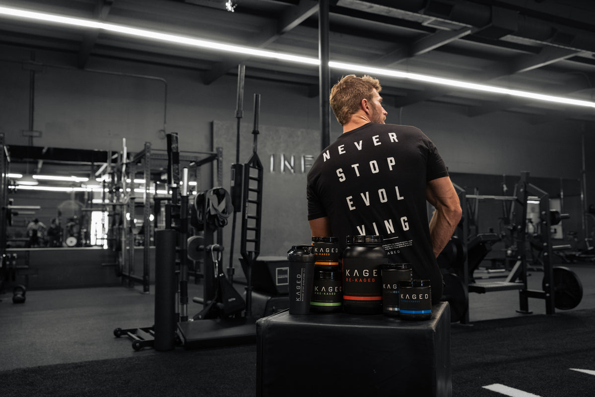 Rear view of black t-shirt on athletic-fit model showing large back print "Never Stop Evolving" in a gym with Kaged supplement containers in the foreground