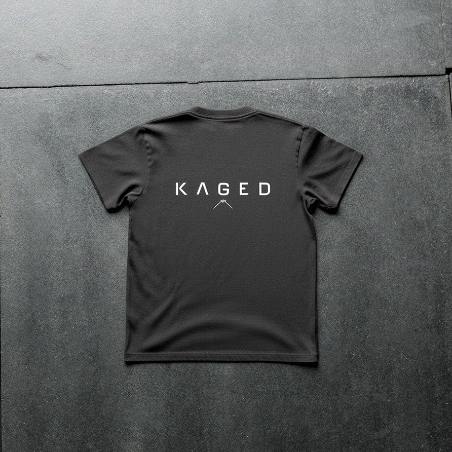 Back view of Ascent Tee showing large white KAGED wordmark and small mountain logo on a black tee against a dark textured floor