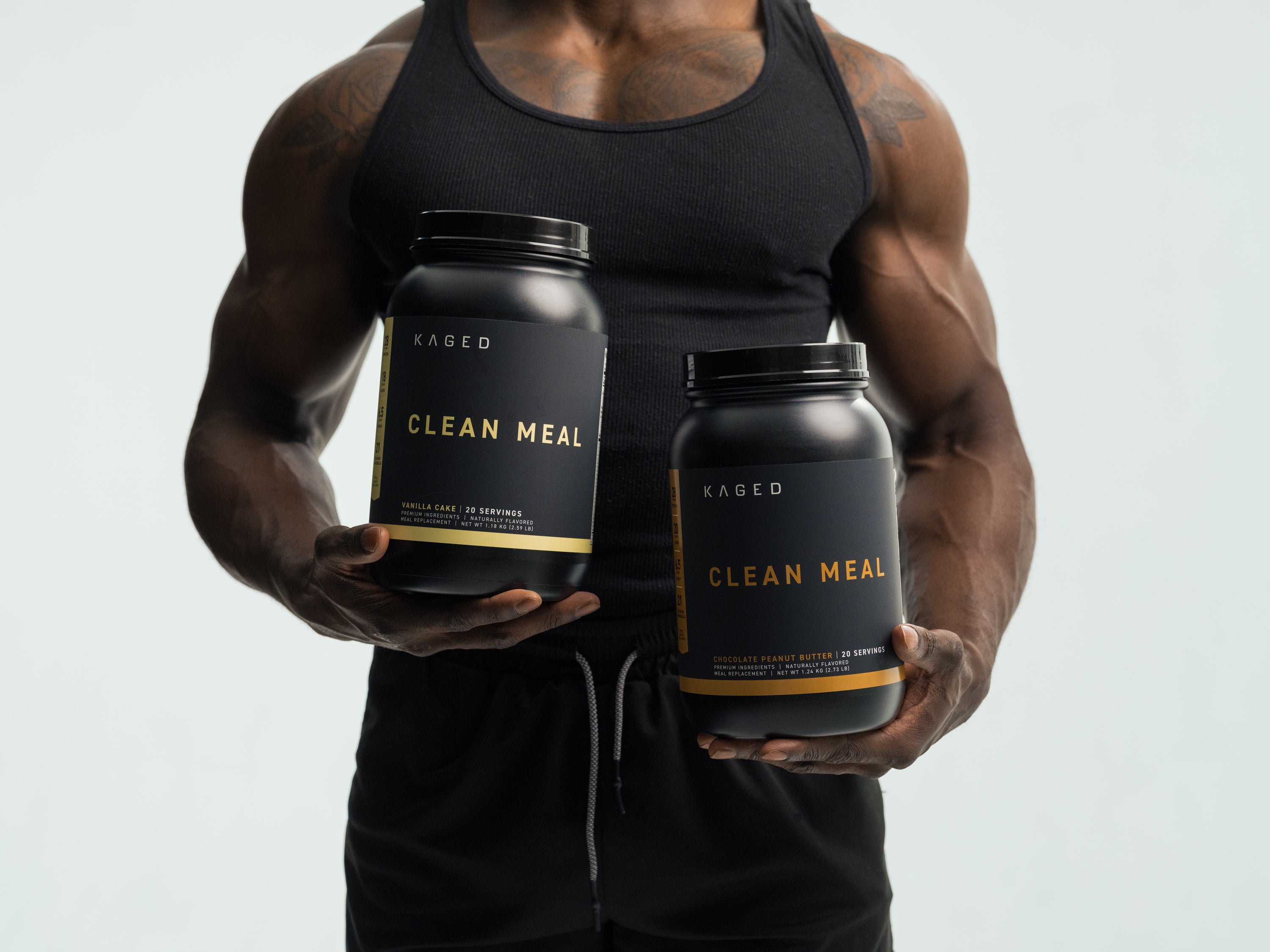Front view of two KAGED Clean Meal meal replacement tubs held by a muscular man, labels reading CLEAN MEAL, light gray background
