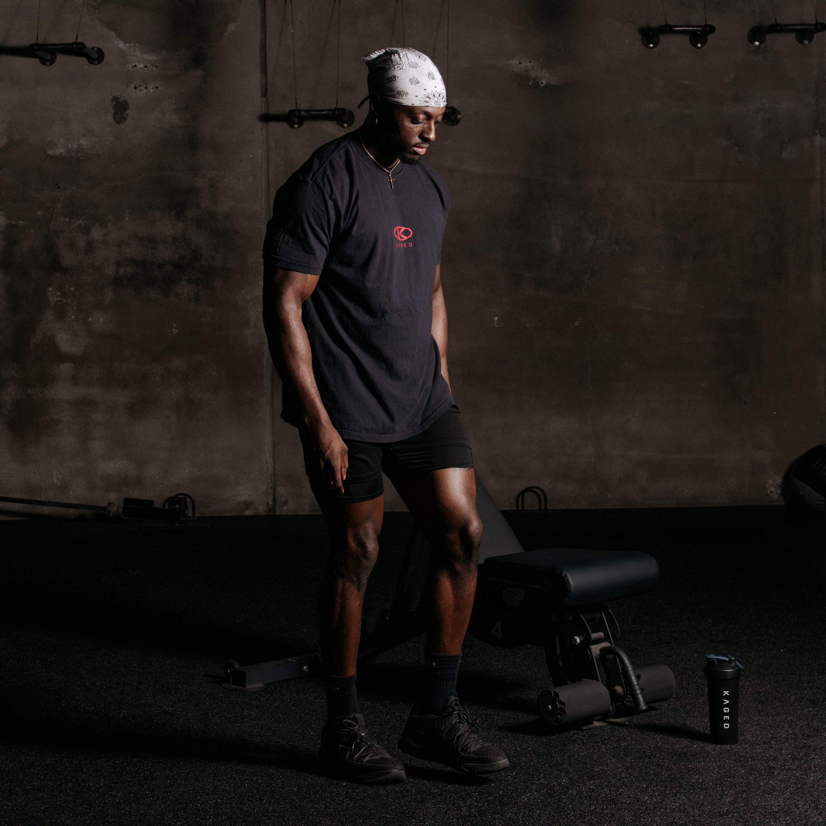 Model wearing black short-sleeve tee with small red Kaged circle logo on chest, standing in a dim gym setting