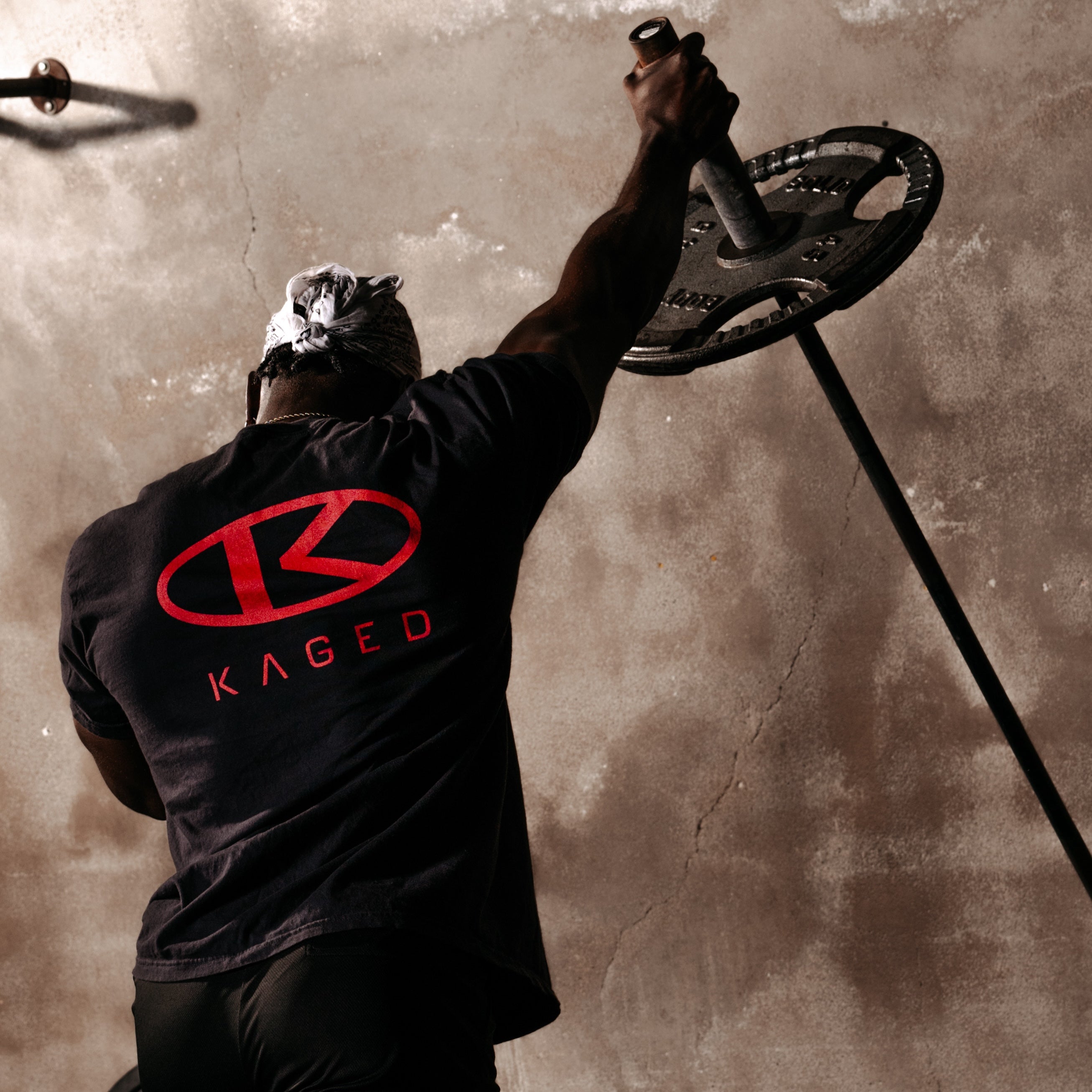 Rear view of Circle Tee showing red KAGED circle logo on the back as a model lifts a barbell against a concrete wall