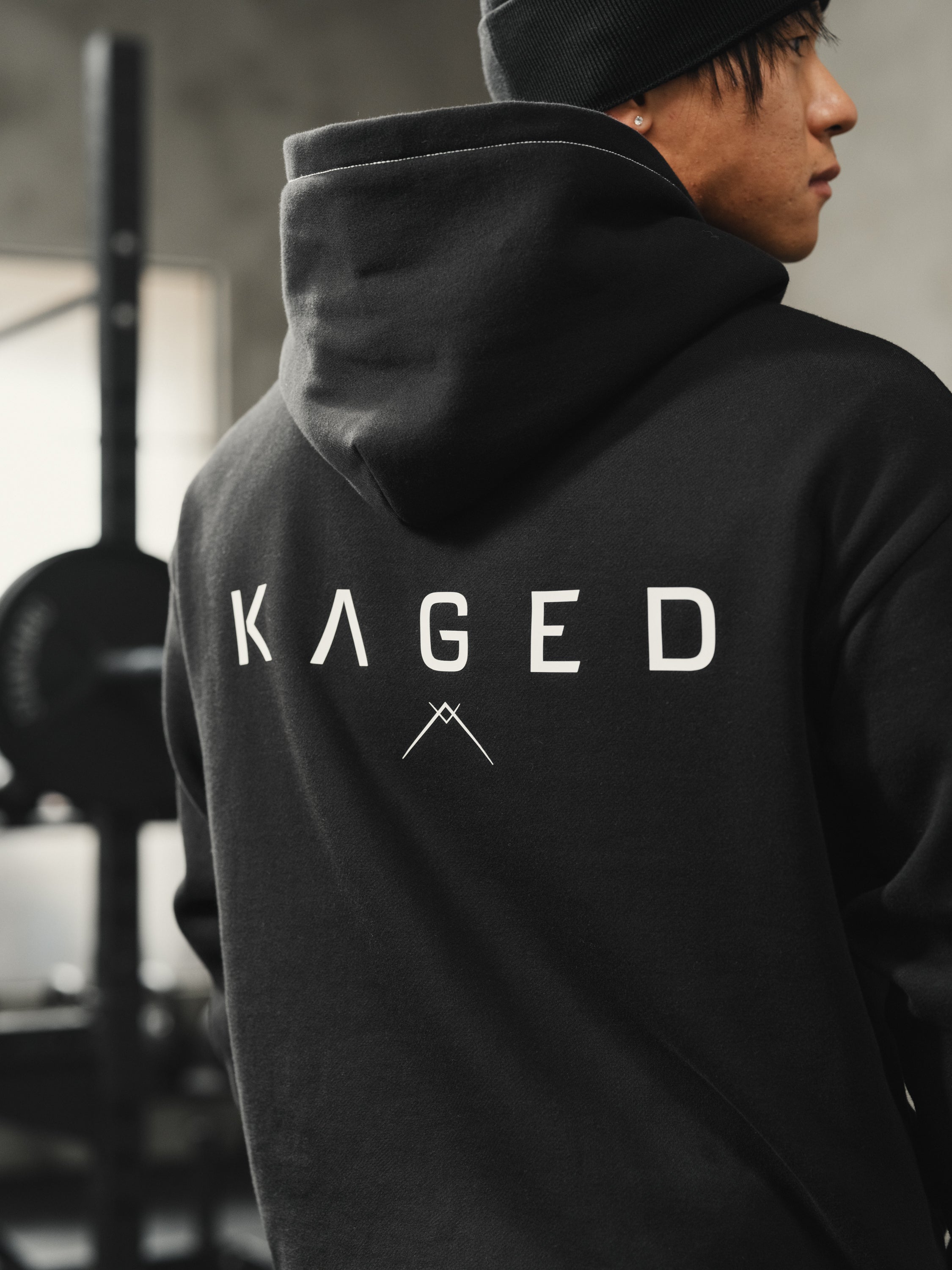 Rear three-quarter view of Ascent Hoodie showcasing white KAGED back print and relaxed athletic fit in a gym