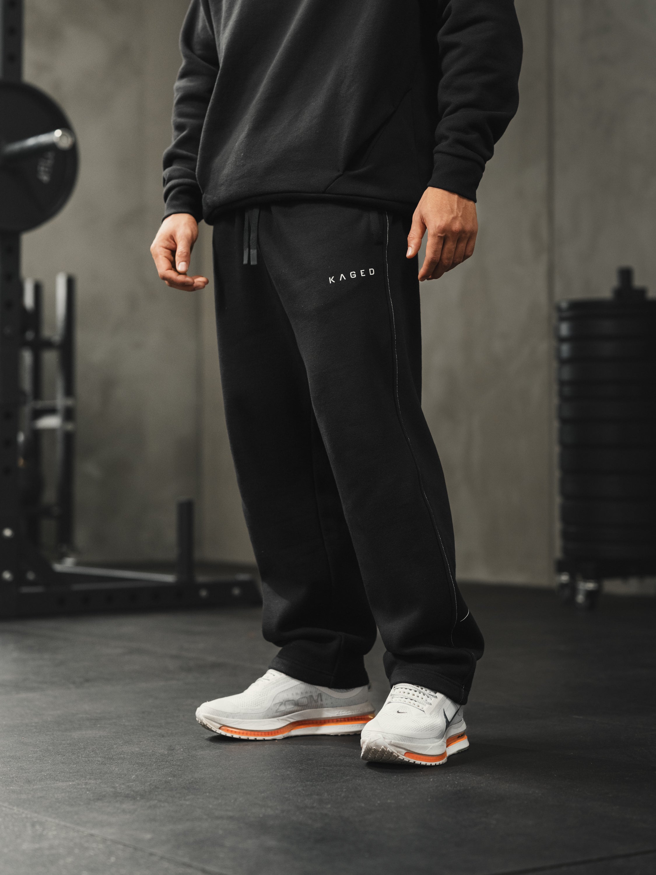 Front view of black Ascent straight-leg sweatpants showing white side-seam stitching, Kaged logo on left thigh, drawstring waistband, gym setting