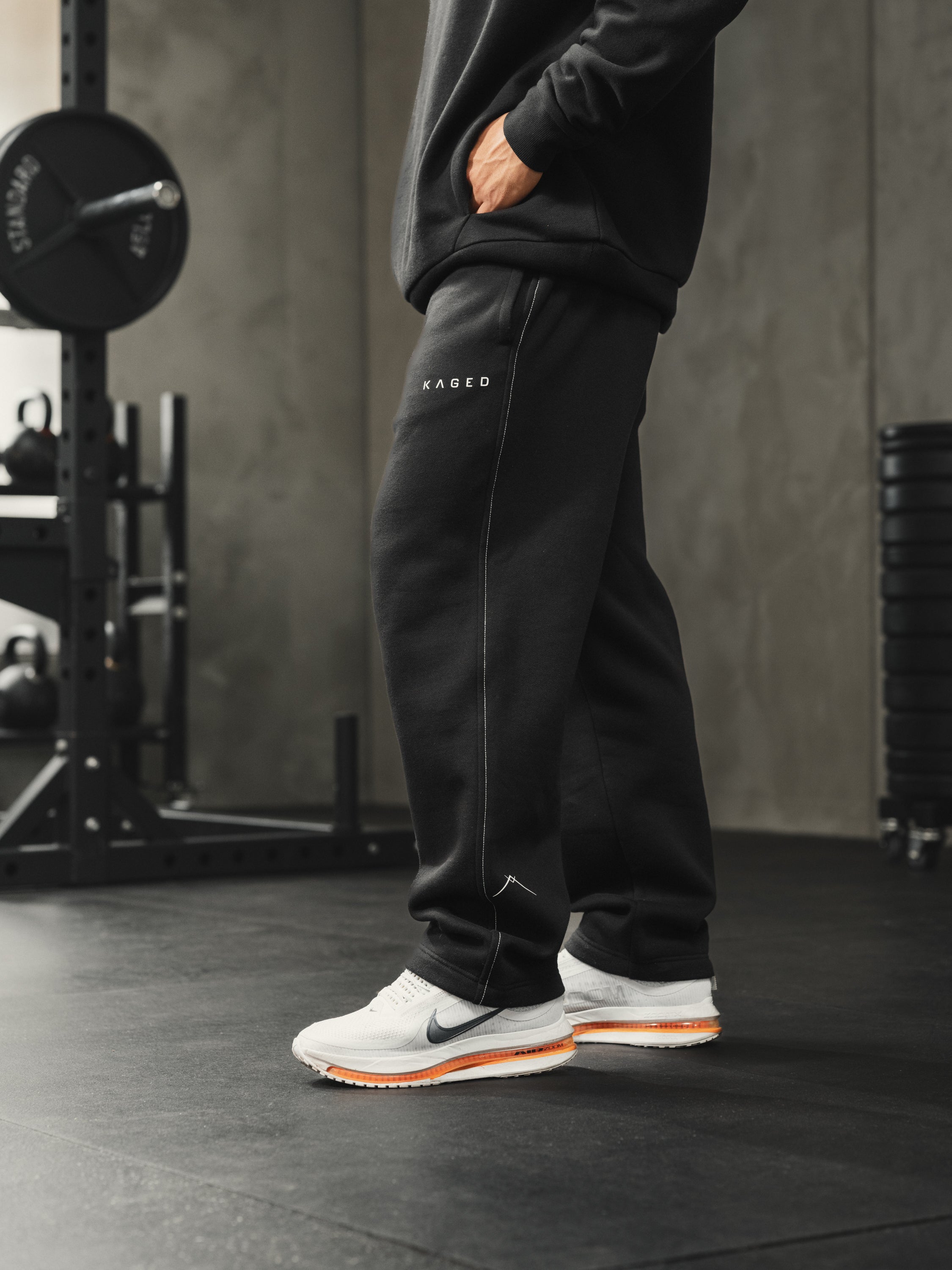 Side view of Ascent straight-leg sweats in black showing white side-seam stitching, KAGED logo and Ascent ankle symbol in a gym setting