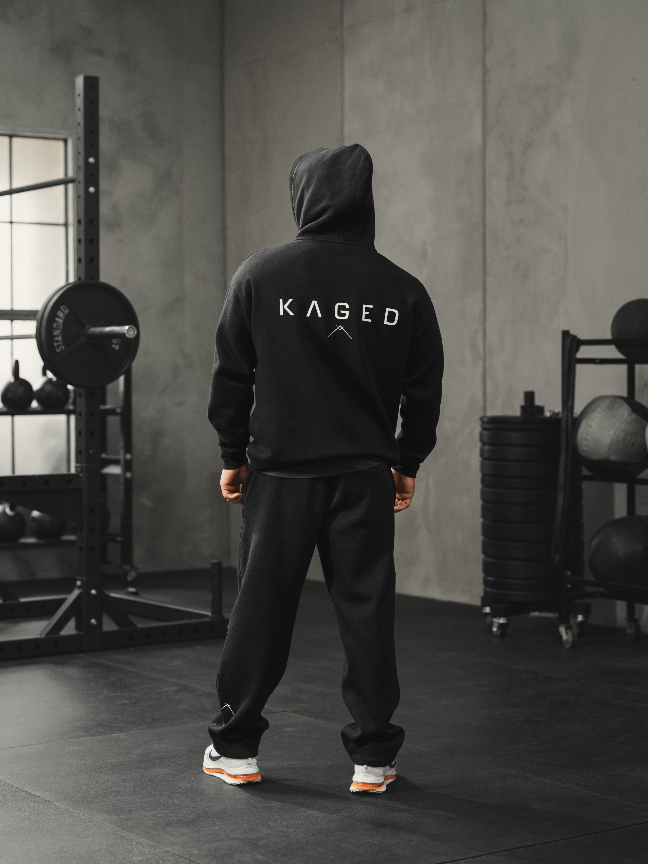 Rear view of Ascent Hoodie with hood up, showing large KAGED logo on back and relaxed athletic fit on model in gym setting