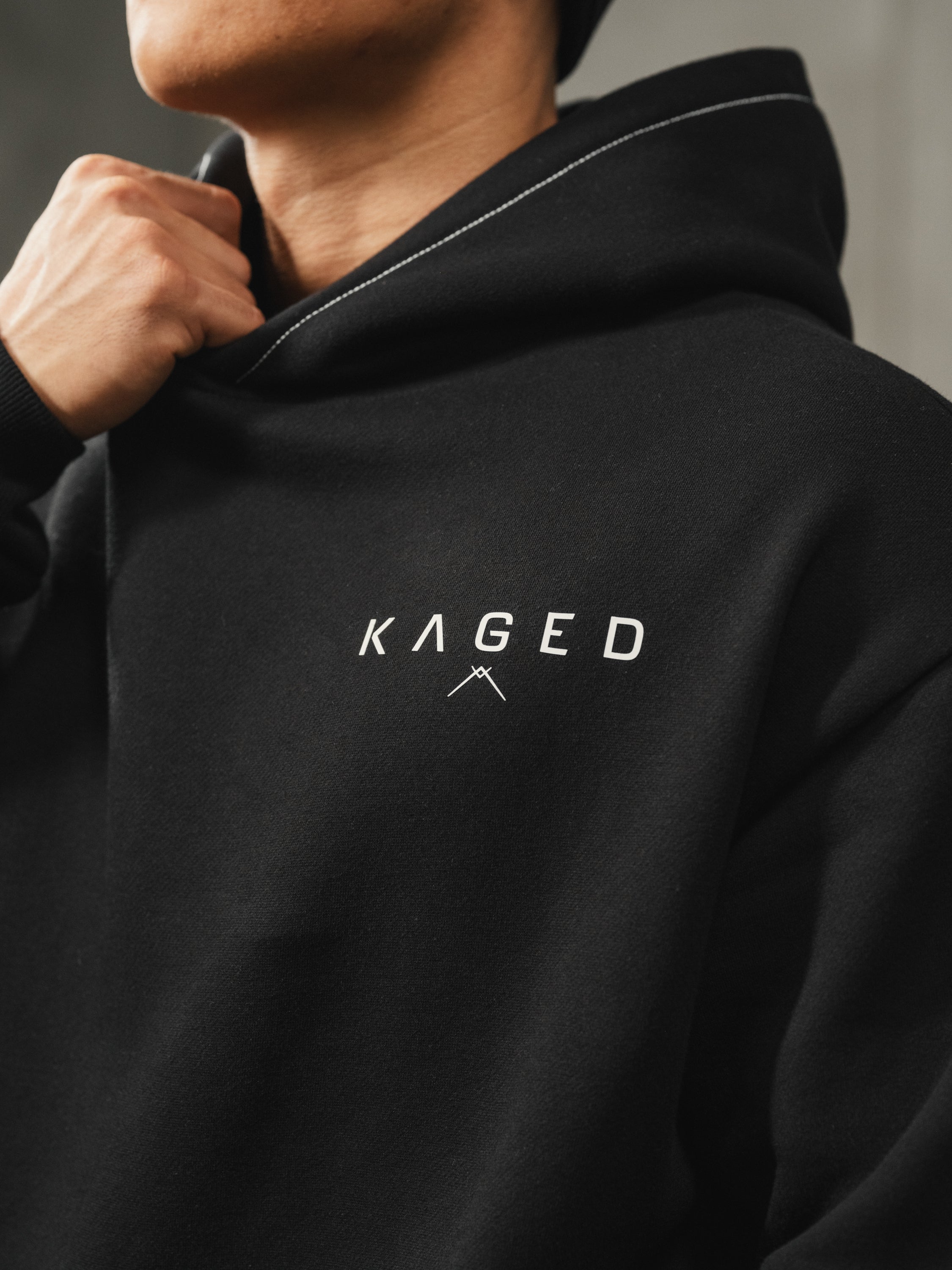 Close-up of Ascent hoodie on model showing white KAGED chest logo, contrast stitching on hood, black color and relaxed athletic fit
