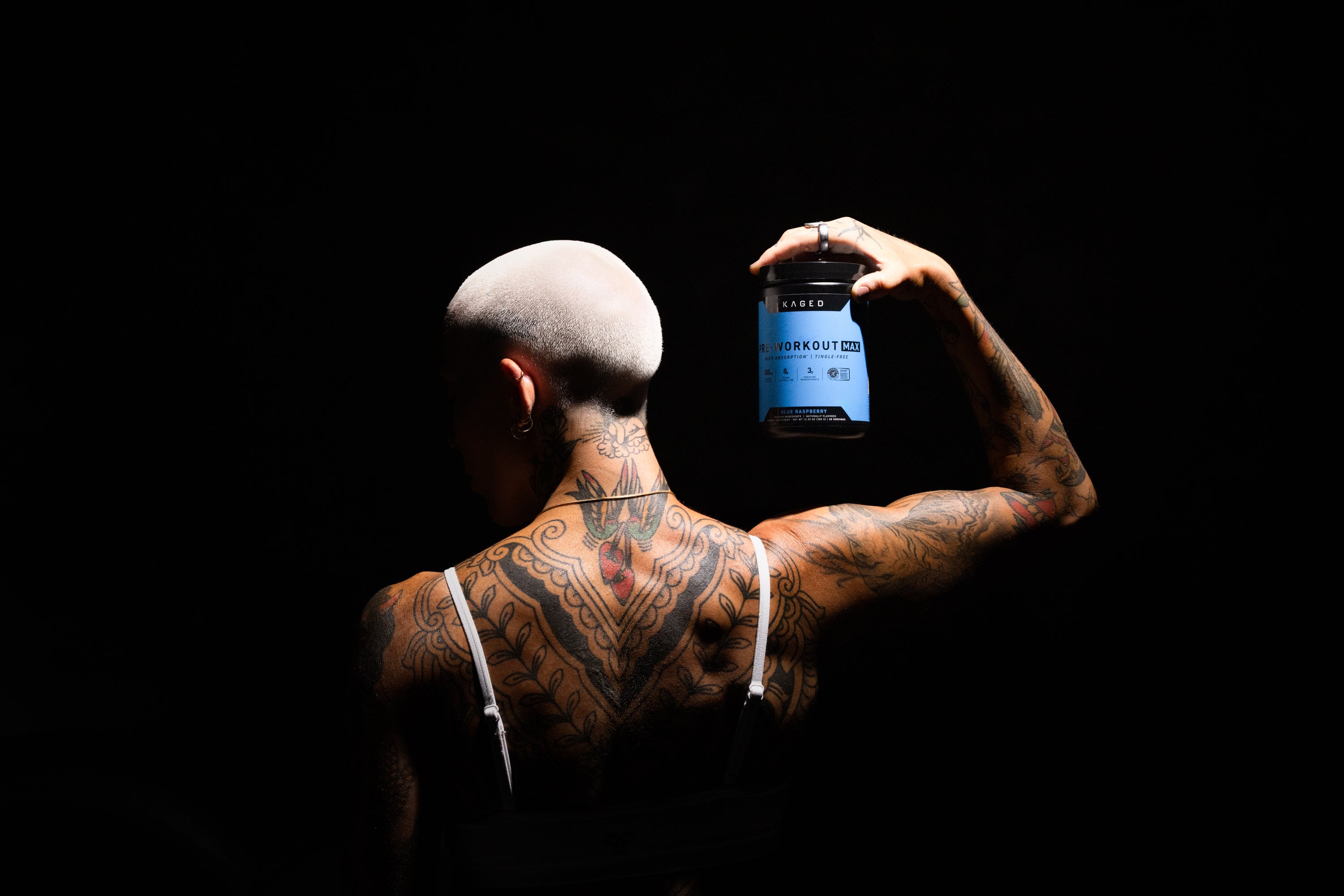 Overhead view of Pre-Workout Max tub showing blue label and black lid, held by tattooed model against dark background